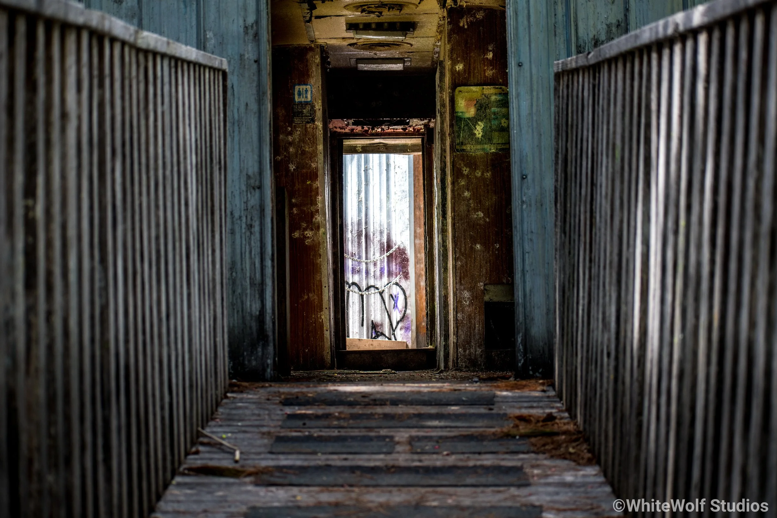 A dingy, abandoned balcony with rusty railings and peeling paint, leading down into a darker area with graffiti on the wall.