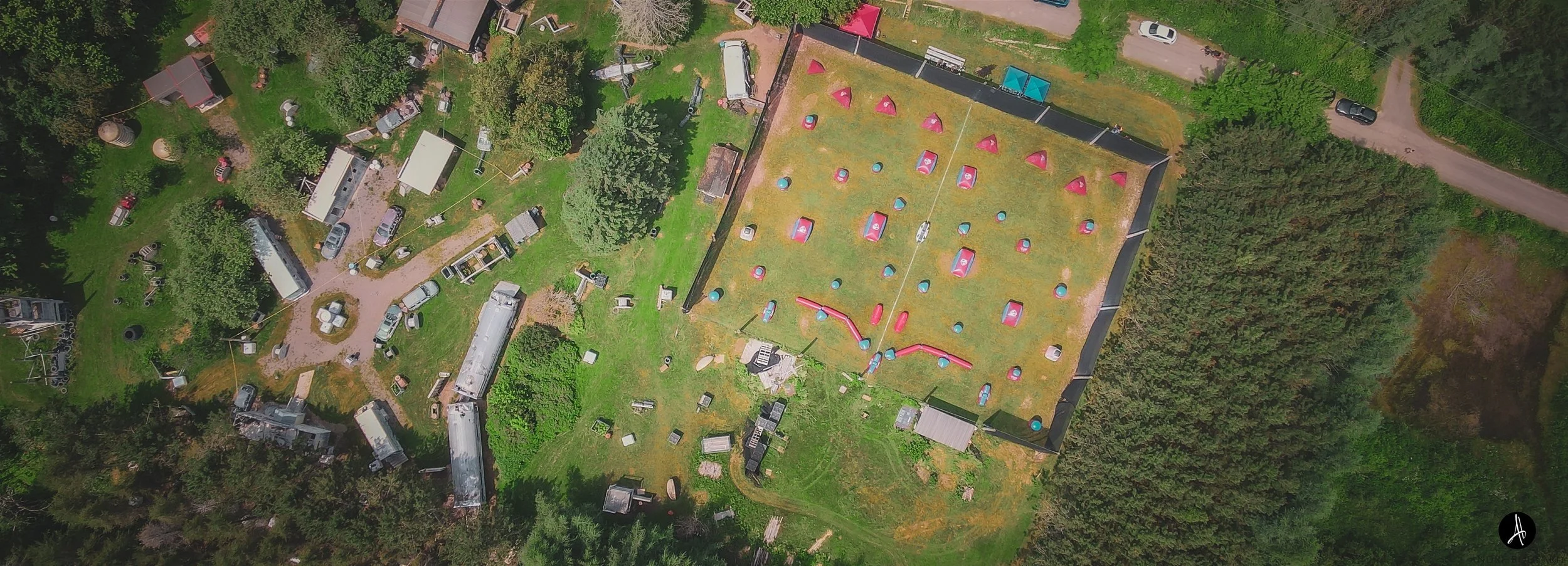 An aerial view of a paintball field surrounded by trees, with several structures, vehicles, and scattered equipment around it.