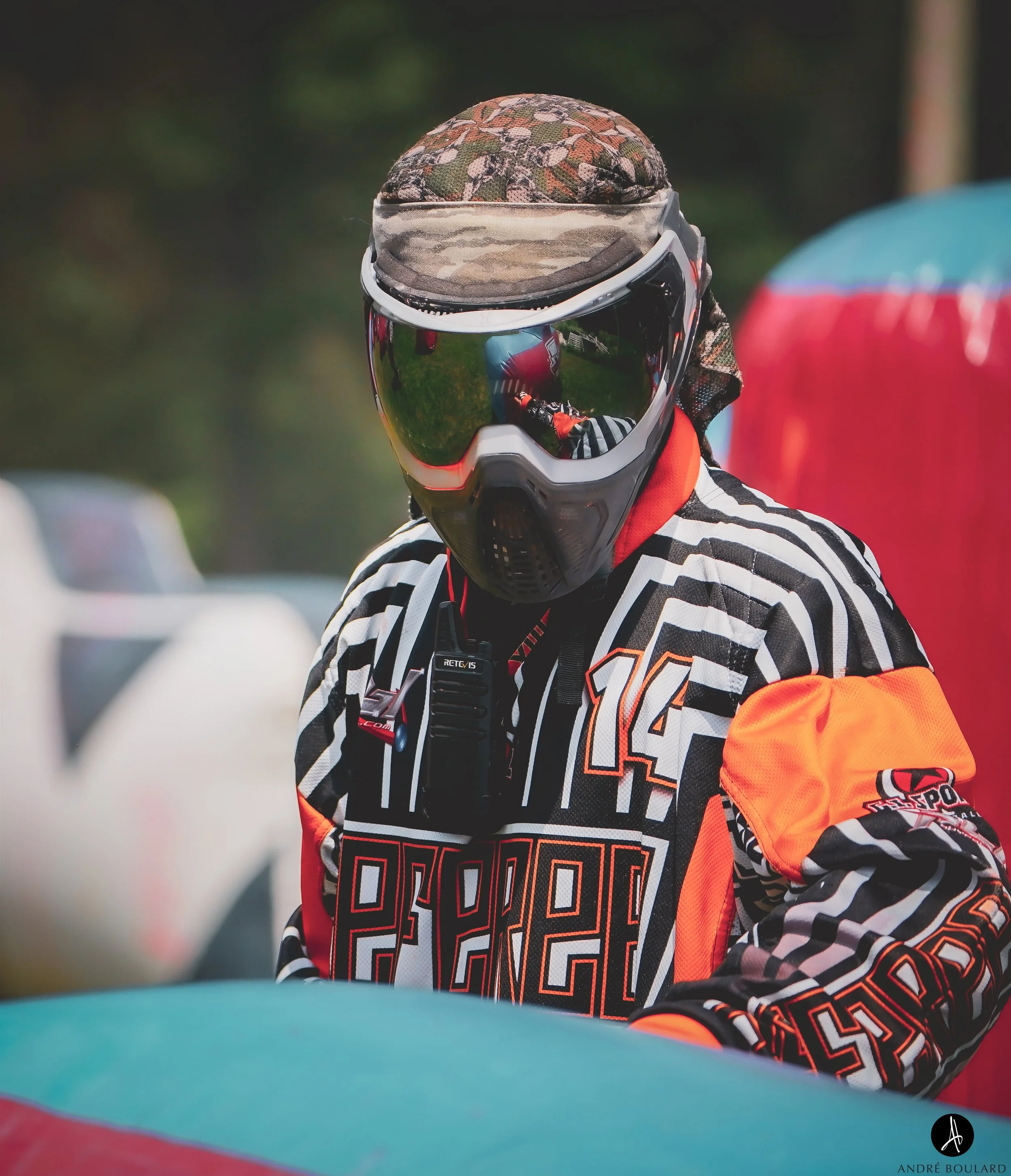A person wearing a black, white, and orange motocross jersey with geometric designs and the number 450, a camouflage hat, and reflective motocross goggles with a camouflage band covering the helmet. The person is outdoors, possibly at a motocross eve