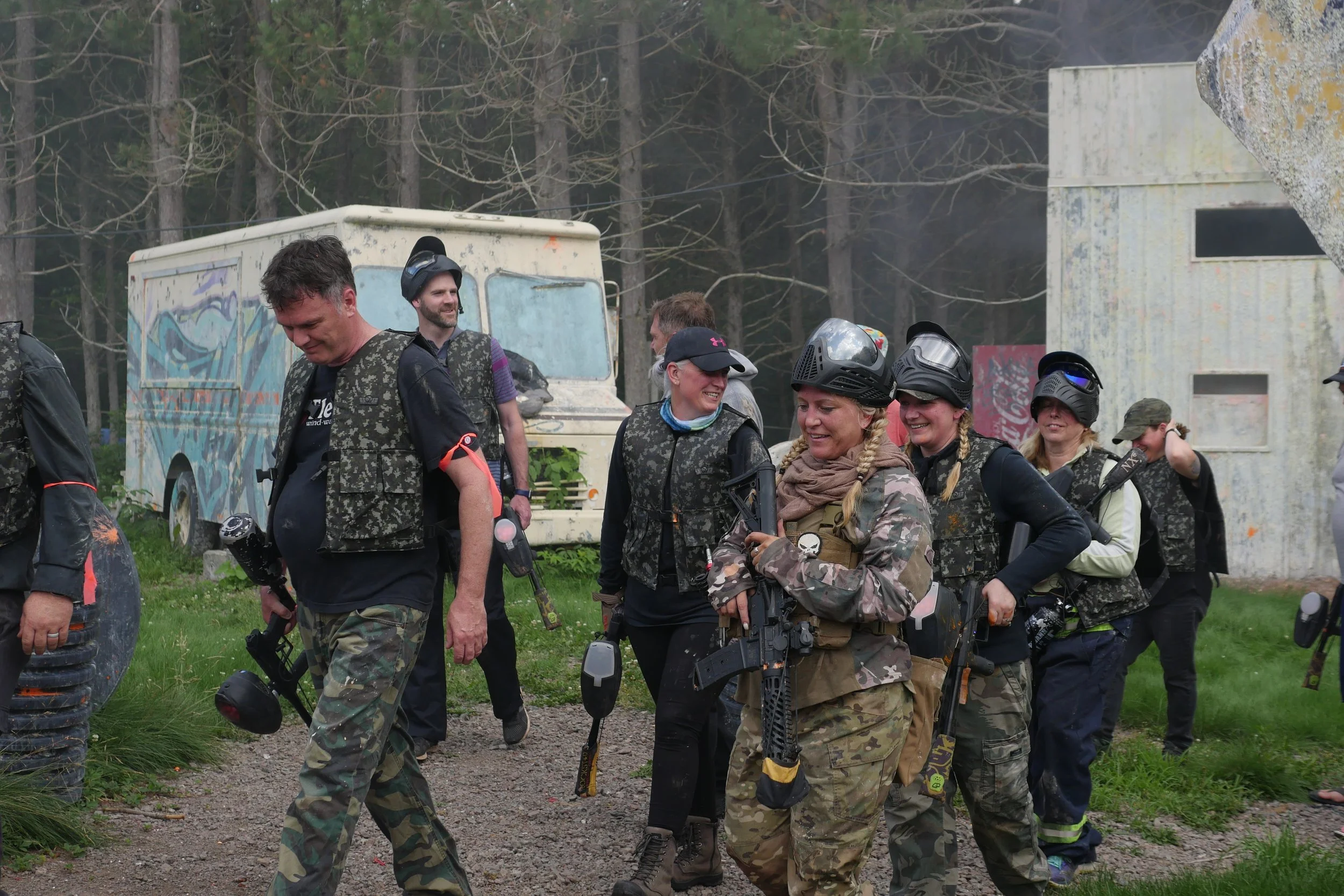 A group of people dressed in camouflage gear, wearing helmets and protective gear, outdoors in a wooded area, smiling and holding paintball guns, with a background of trees, graffiti-covered trailer, and a building.
