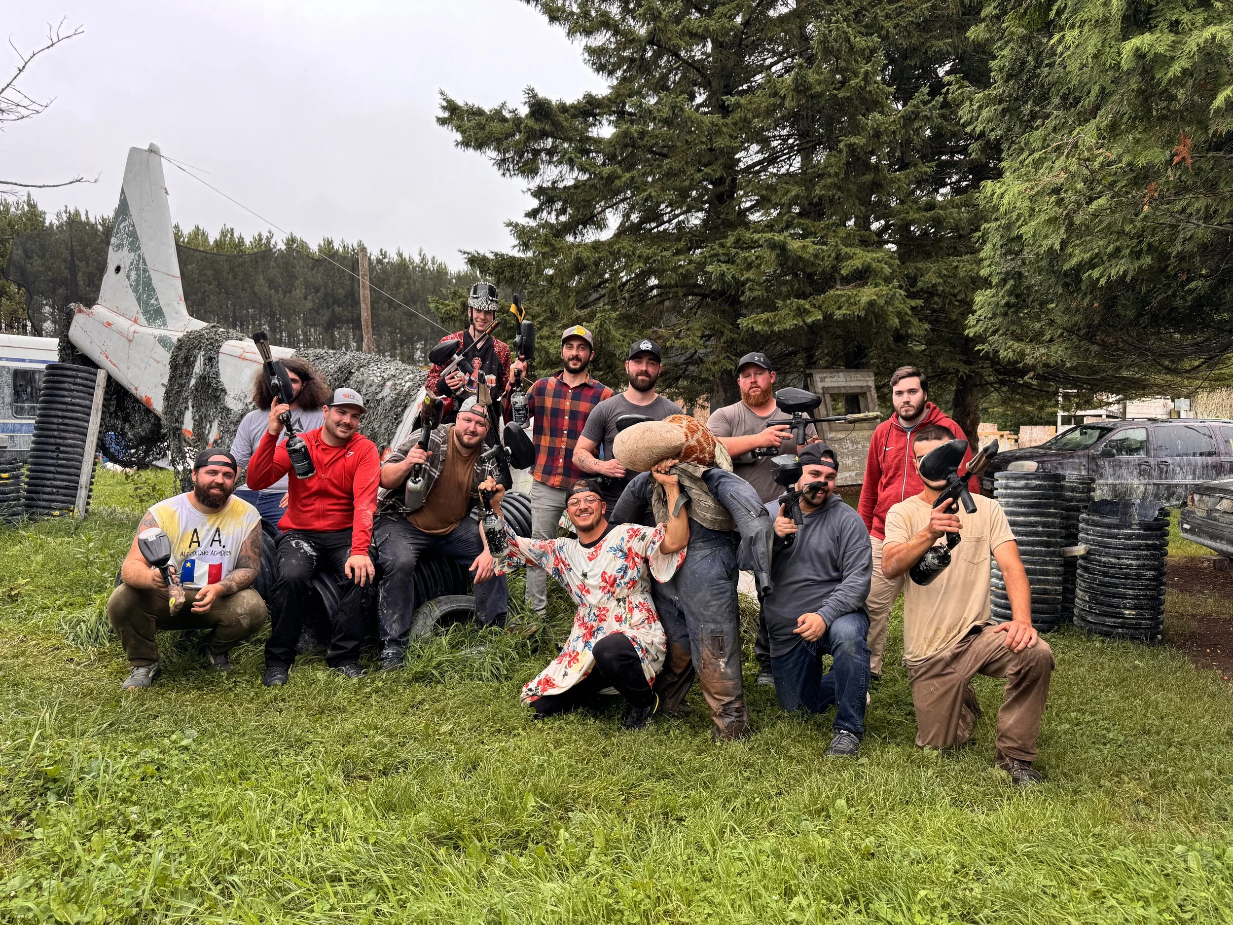 Group of people holding paintball guns, posing outdoors near a fallen airplane, with trees and vehicles in the background.