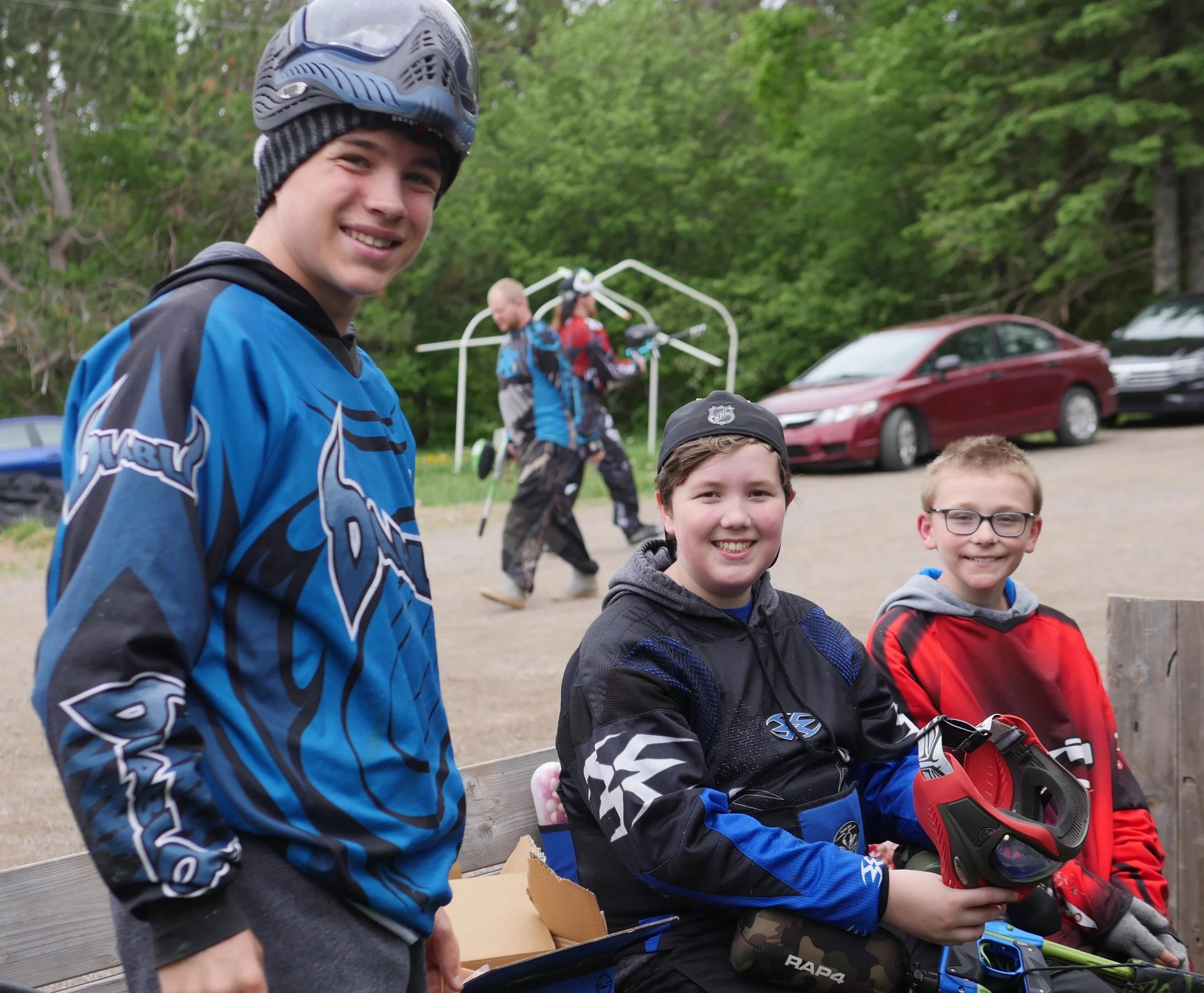 Group of children at outdoor event, some wearing hockey gear, in a natural setting with trees and parked cars in the background.
