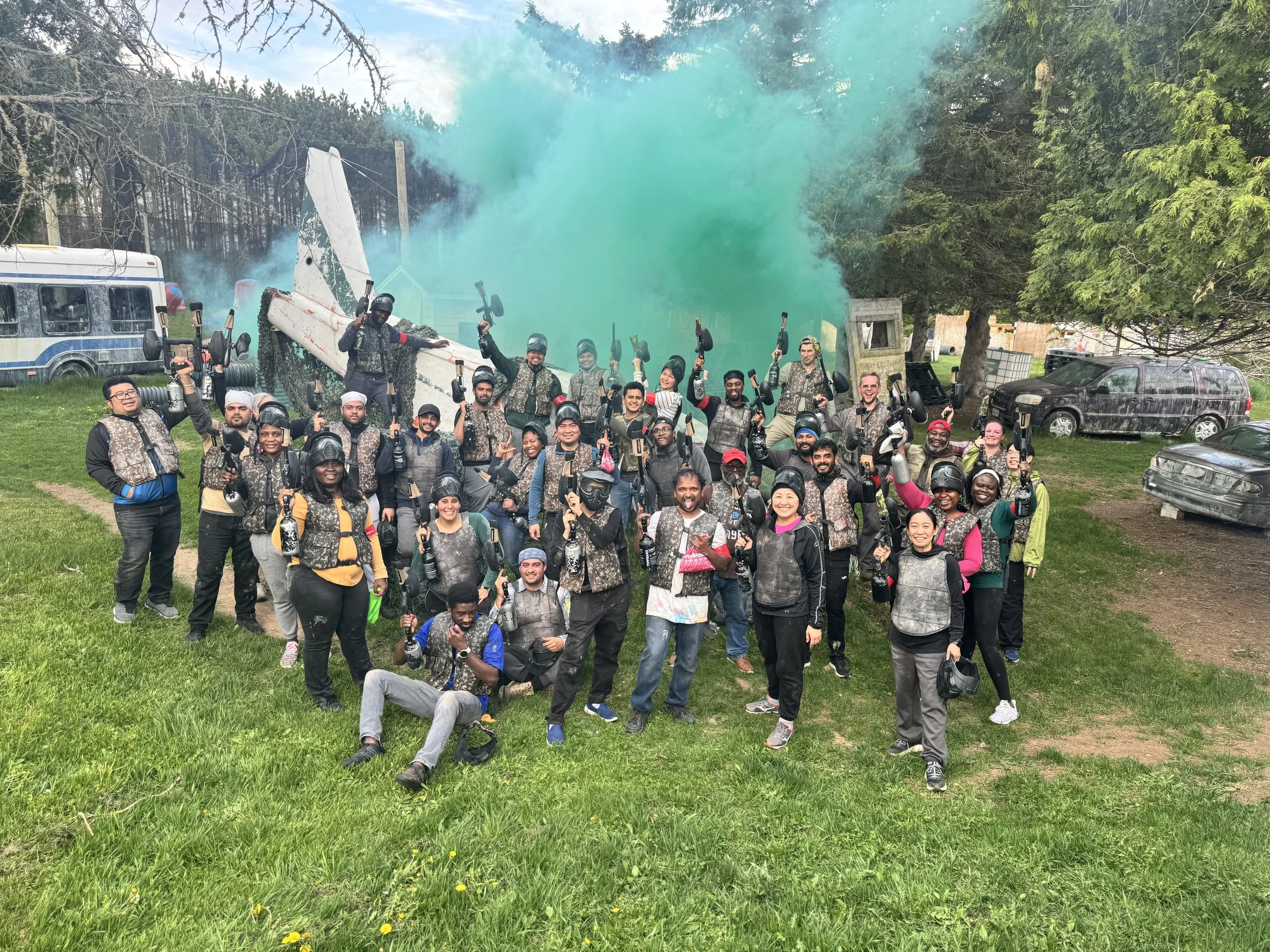 Group of people outdoors holding paintball guns, some wearing helmets, with green smoke in the background, surrounded by trees and parked vehicles.
