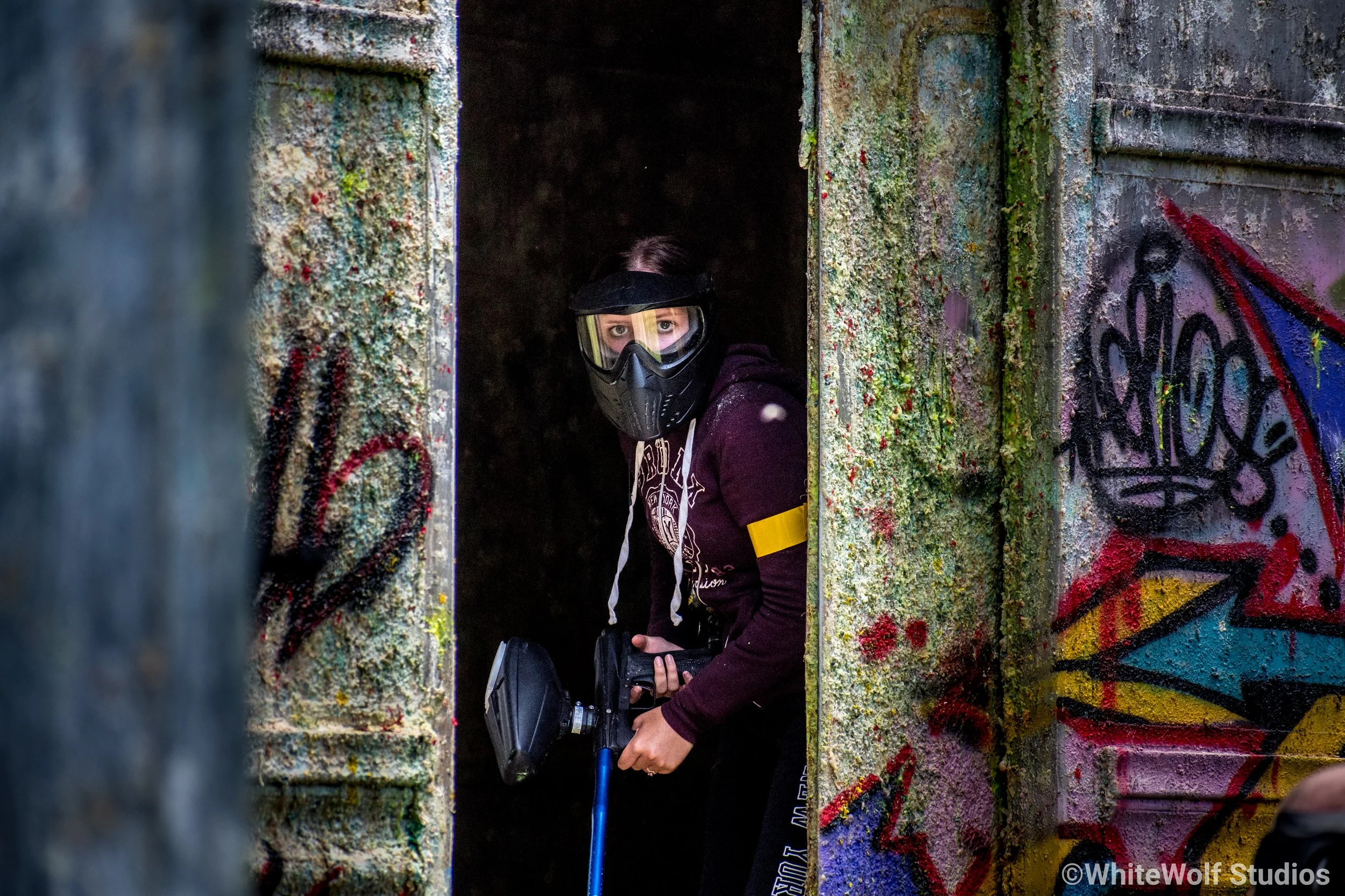 A person wearing a paintball mask and holding a paintball gun peeks through an opening in a graffiti-covered concrete structure.