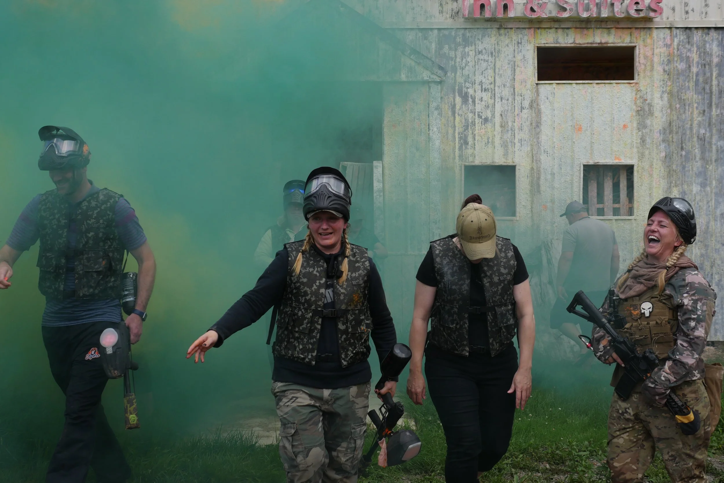 Group of people wearing paintball gear, helmets, and camouflage clothing, standing outdoors amidst green smoke with a weathered building in the background.