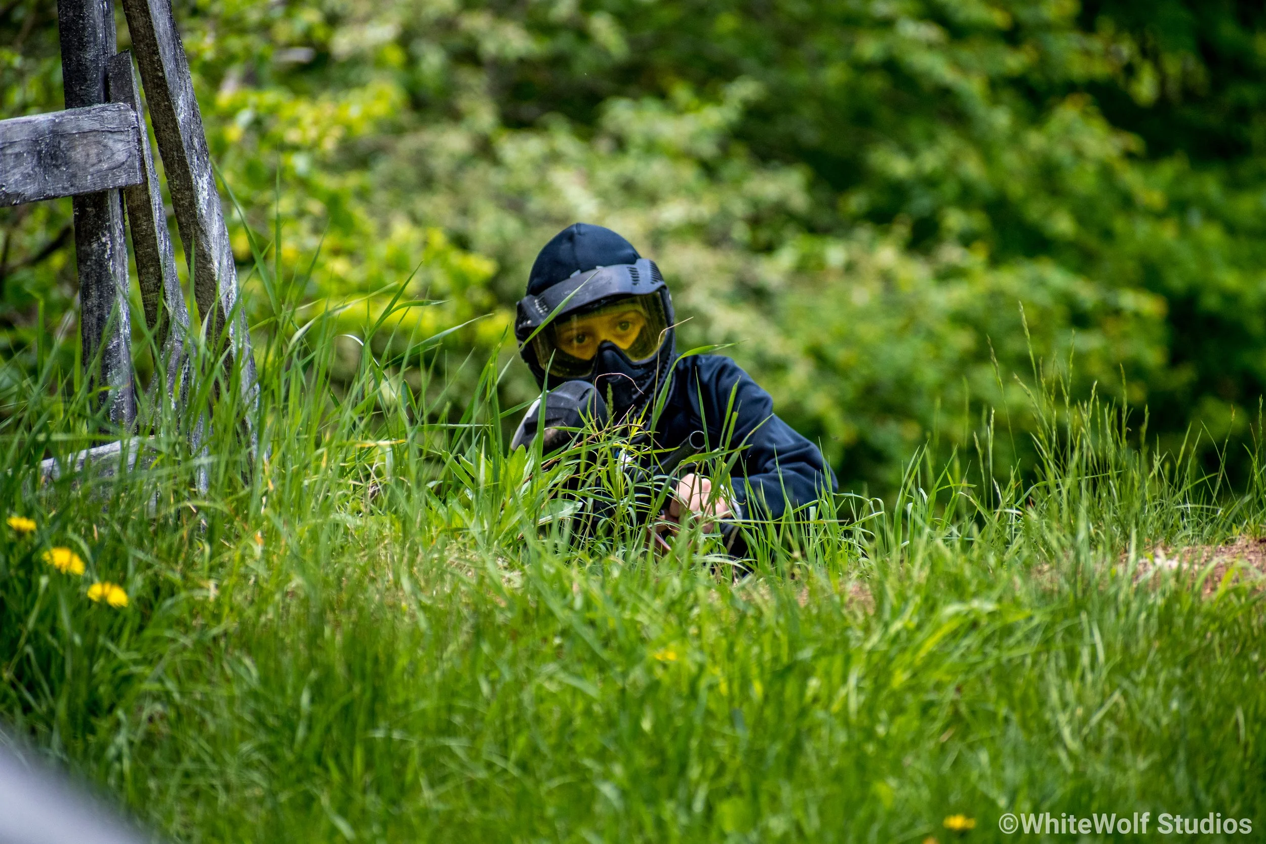 A person wearing a helmet and goggles crouching among tall grass in a green outdoor setting.