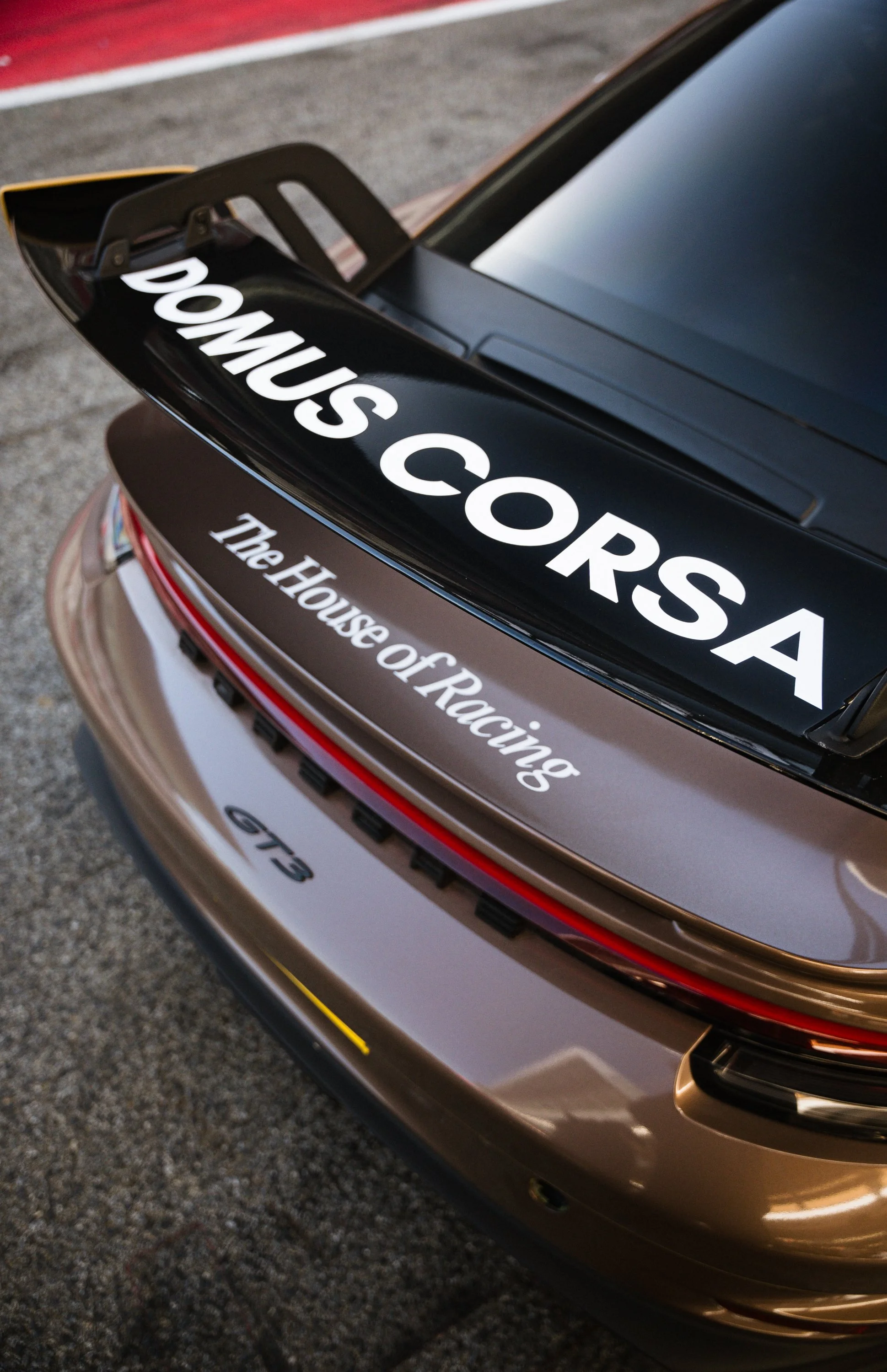 Close-up of the rear of a race car, showing a black rear wing with white text that reads "DONUS CORS..." and a brownish metallic body with the logo "GT3." The car is parked on a textured asphalt surface.