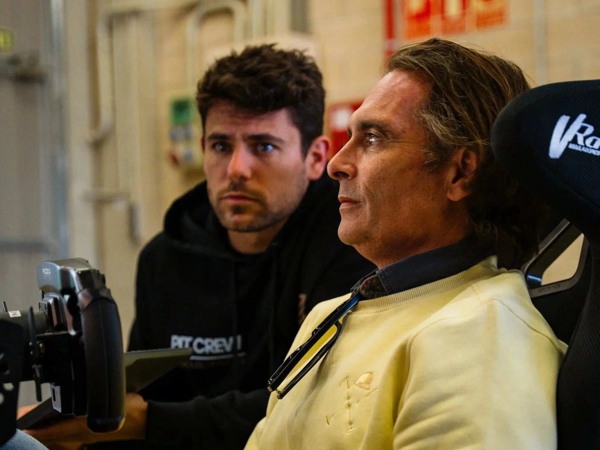 Two men are sitting and looking intently at a device or screen in front of them, one wearing a yellow hoodie and the other in a black hoodie with some text on it. The background is a room with posters and equipment.