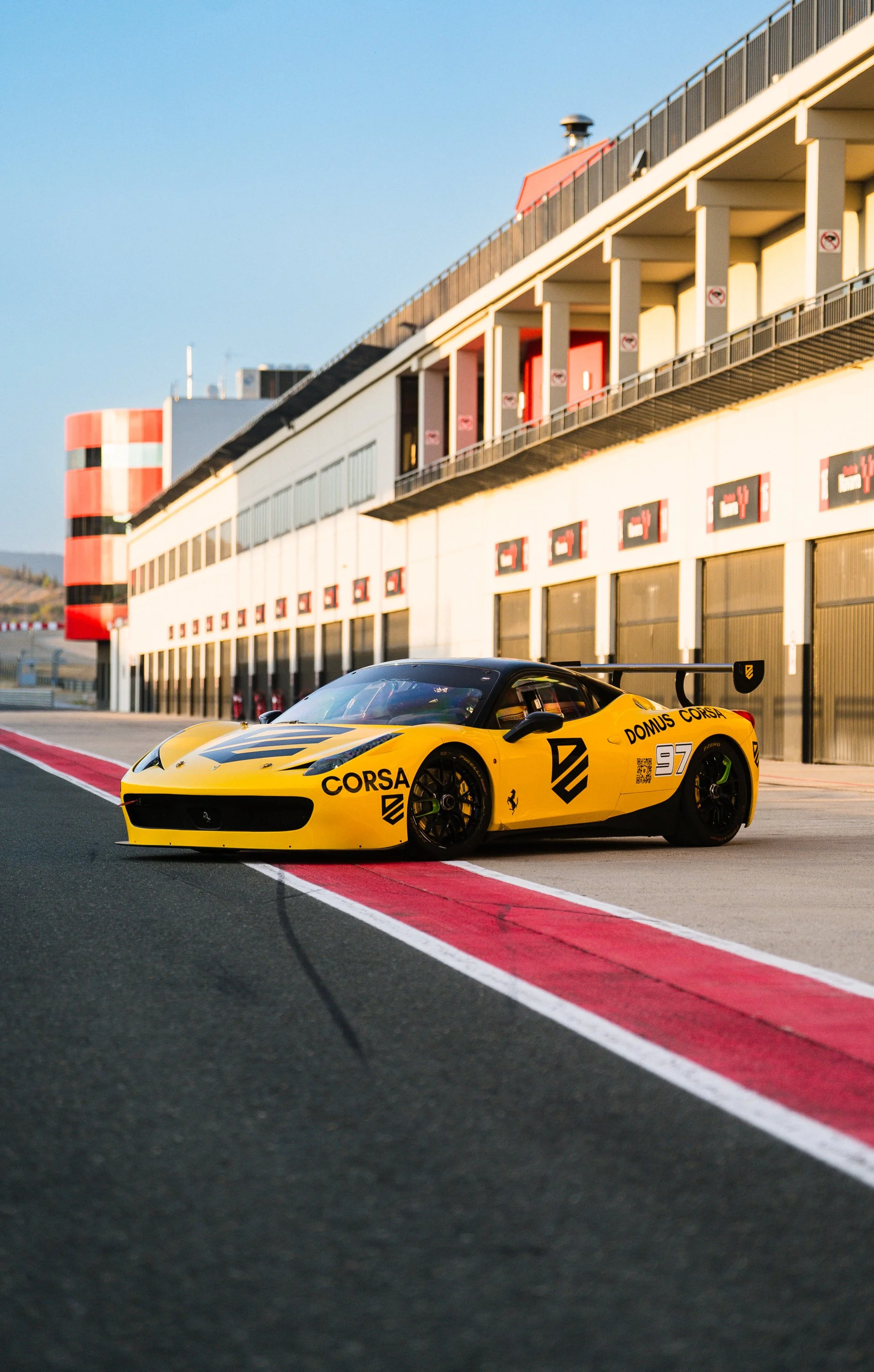 Yellow race car with sponsor logos parked on a racing track near a garage building with multiple signs and red and white barriers.