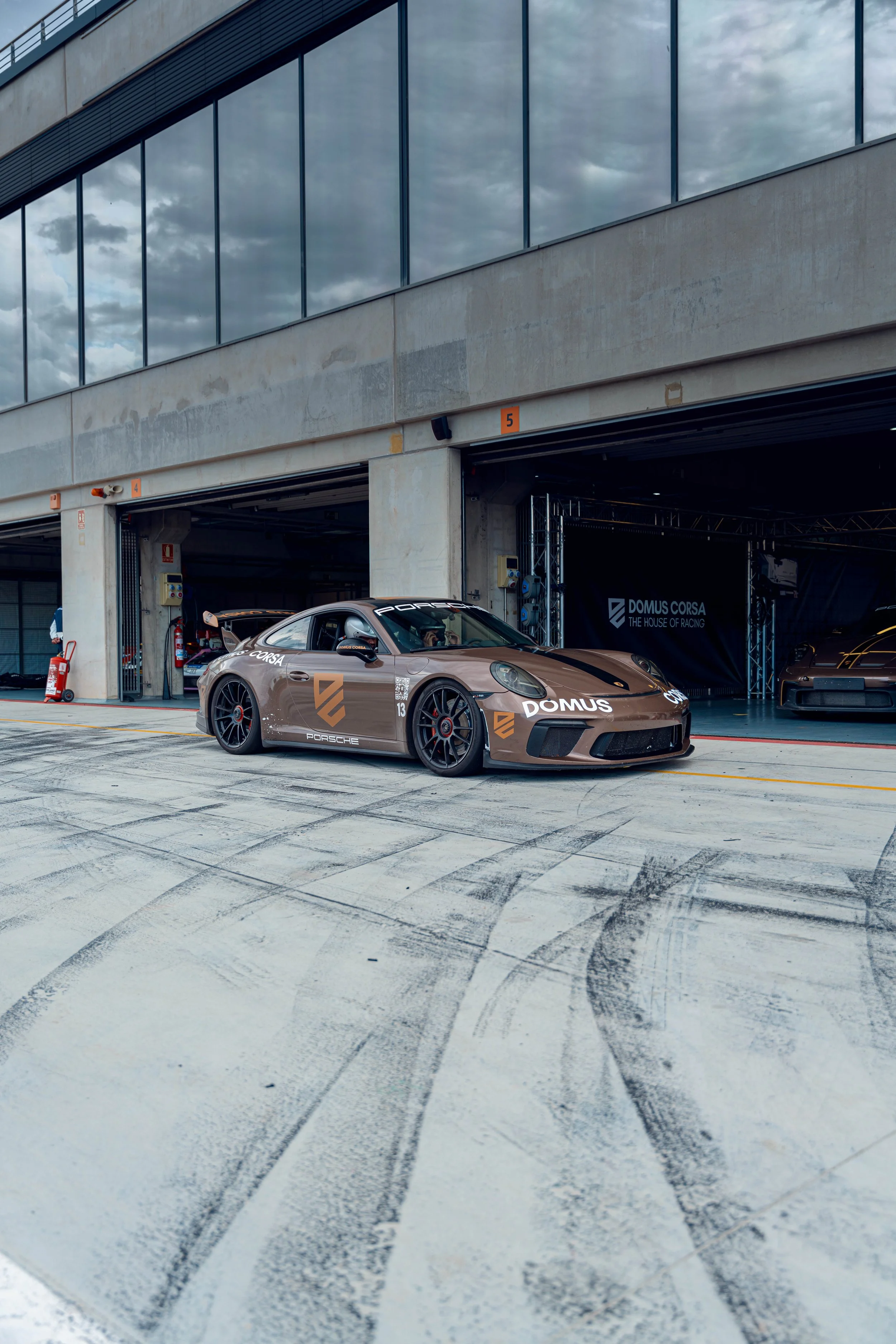 A beige Porsche race car with black and orange decals, number 13, parked in a pit garage at a racing circuit.