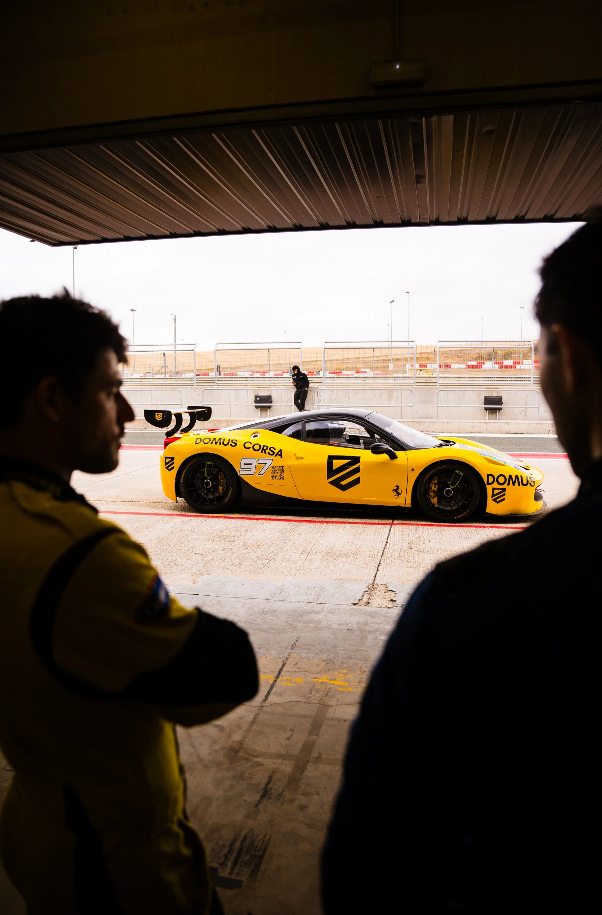 Two race car drivers stand in a garage, looking at a yellow race car with the number 97 on it, at a race track on a cloudy day.