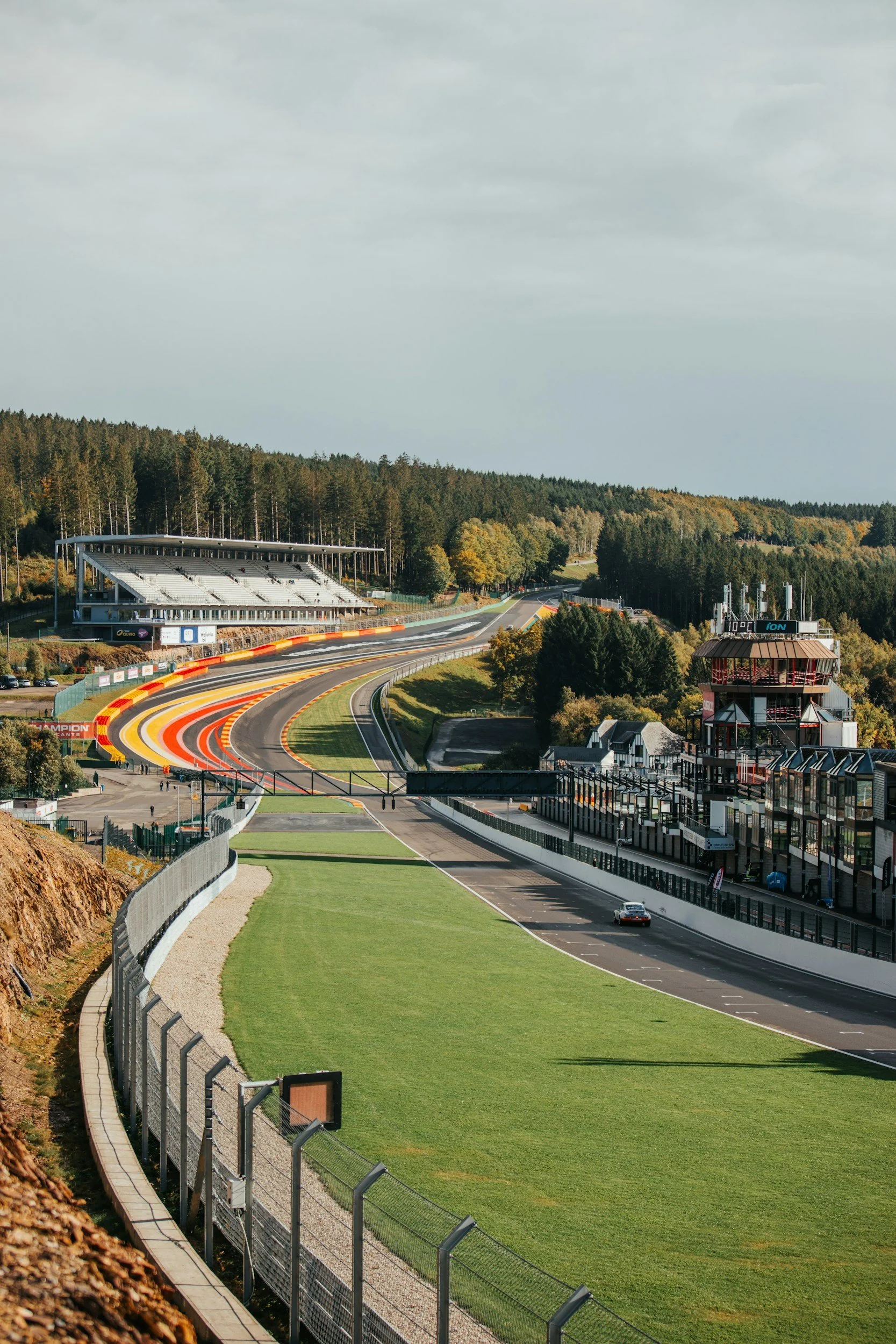 A race track with colorful curbs, grandstands, and a large green grassy area. There are trees in the background and a cloudy sky.