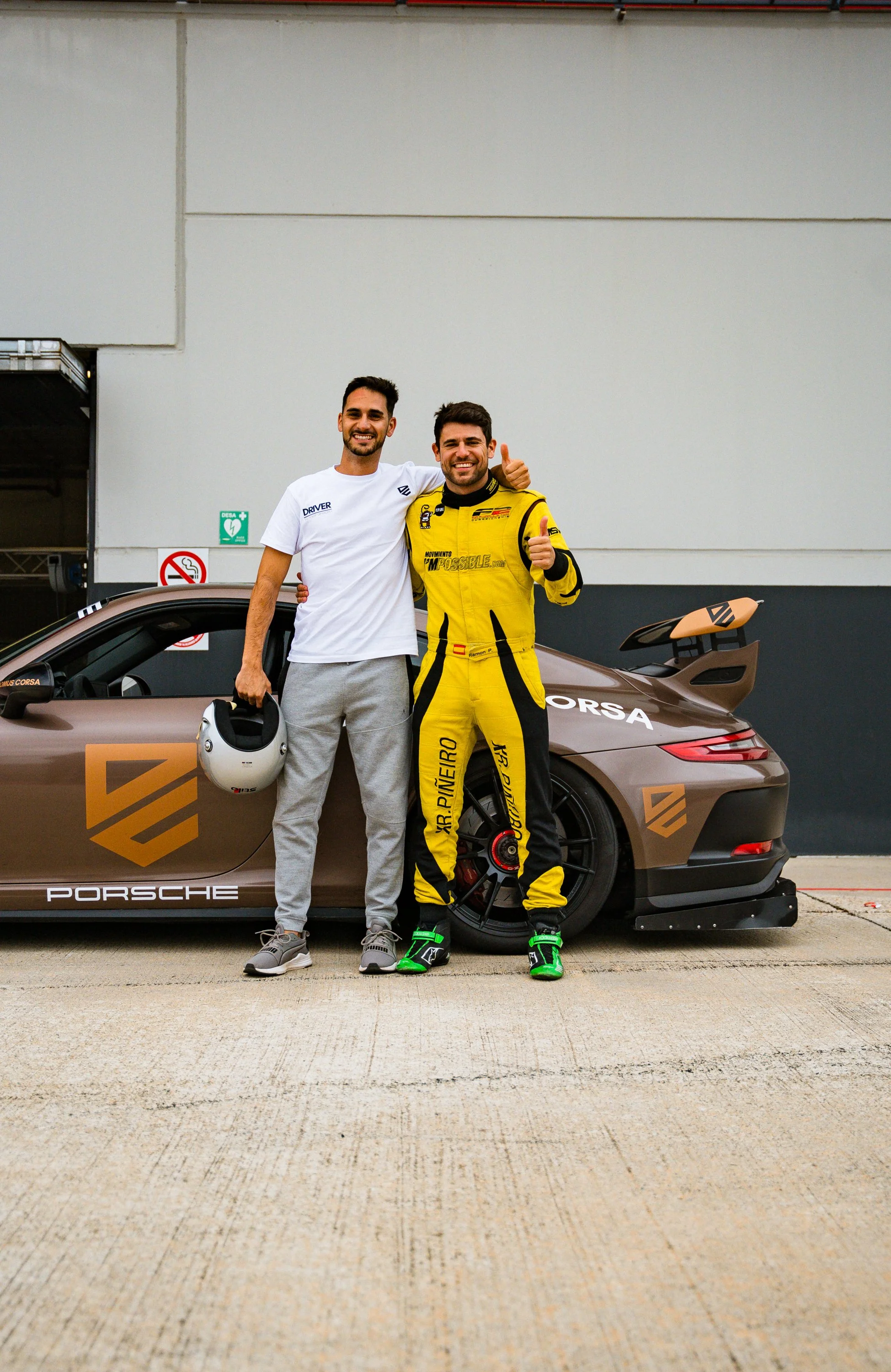Two men standing in front of a brown Porsche race car, one in a white t-shirt and gray pants holding a white helmet, the other in a yellow racing suit with green racing shoes, both smiling and giving thumbs up.