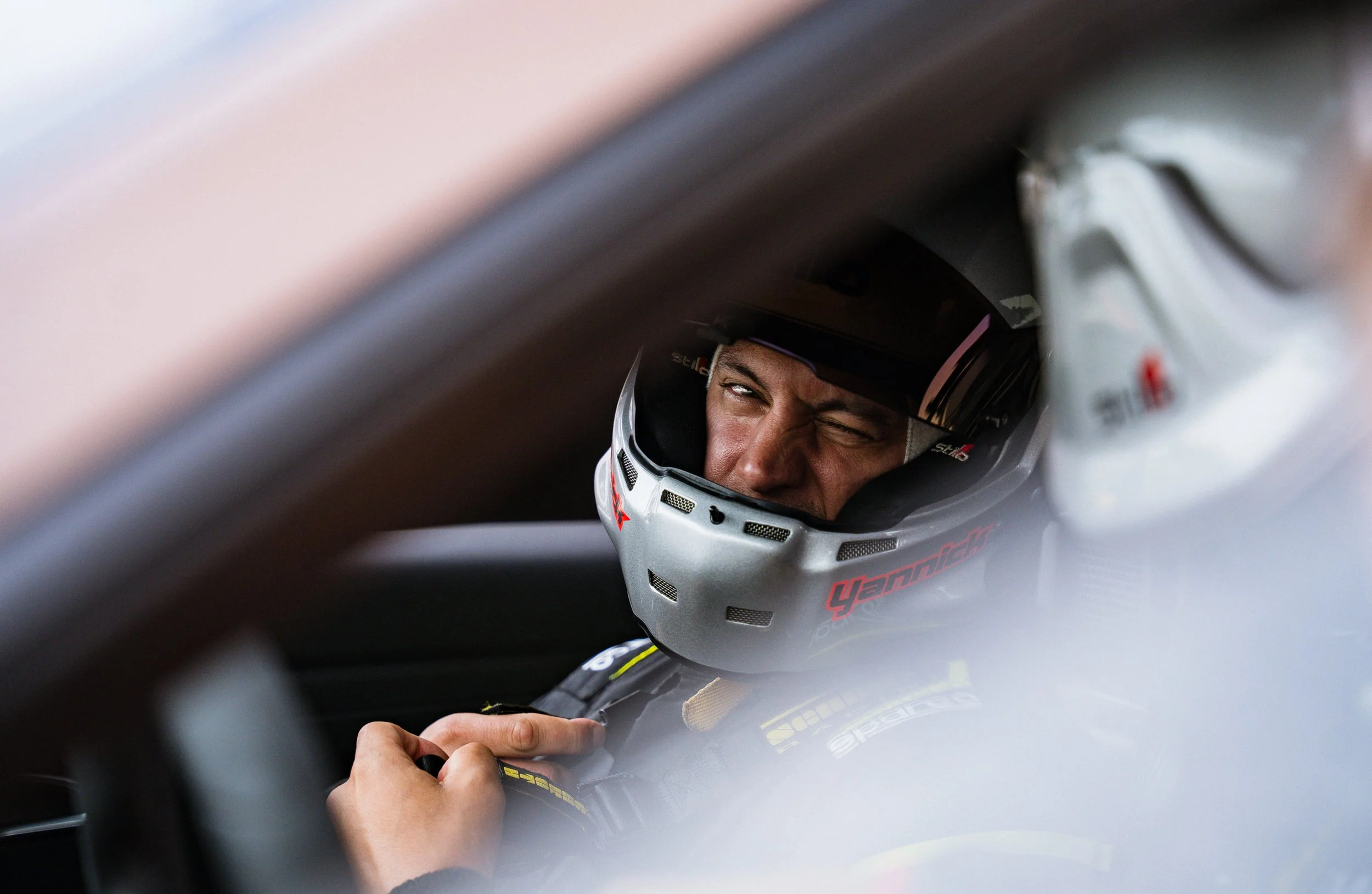 Close-up of a race car driver inside the cockpit, wearing a helmet and showing a focused expression, with someone adjusting his gear.
