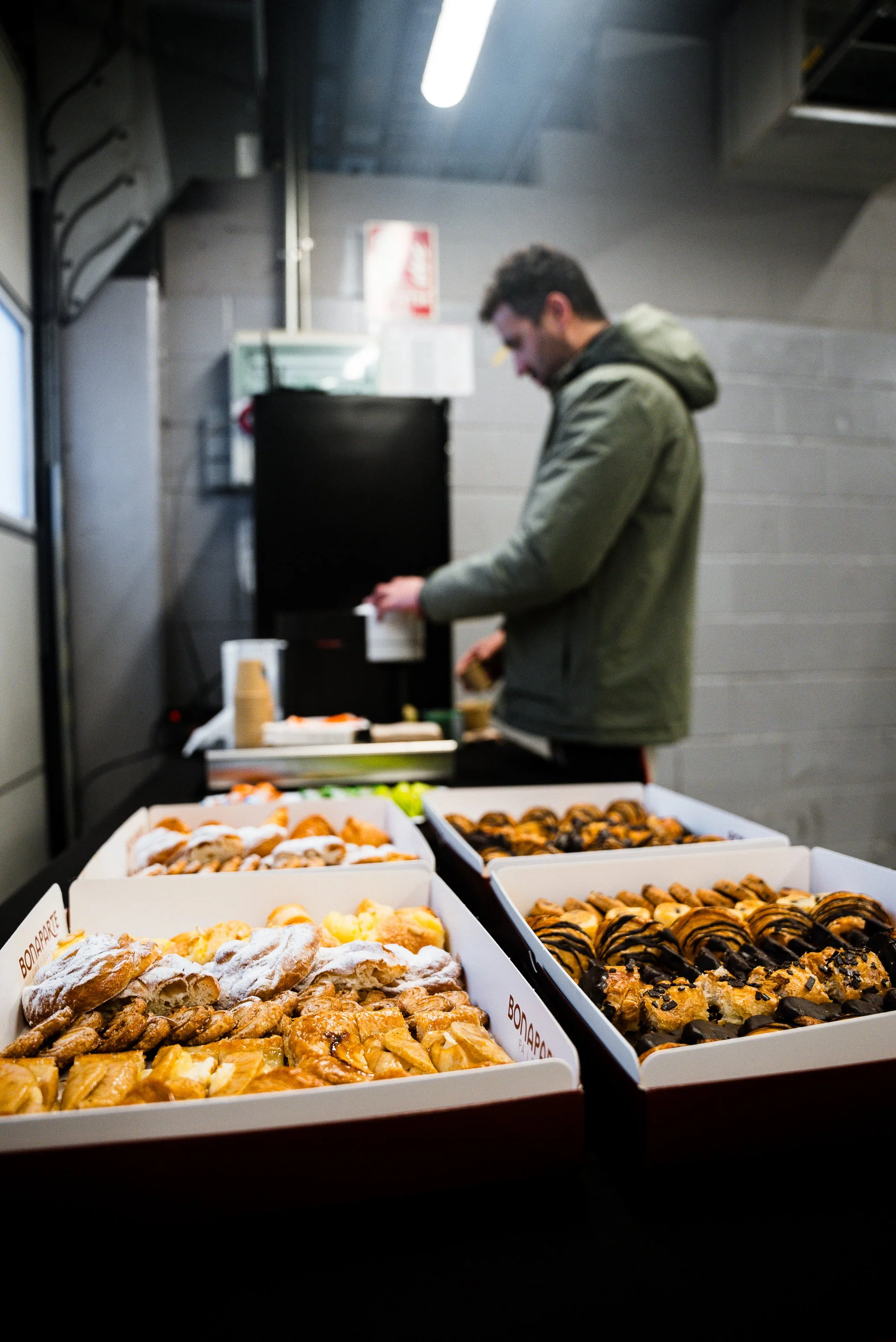 A man in a gray jacket and hoodie stands at a table with various pastries, including croissants, sweet rolls, and chocolate pastries, in a commercial or industrial setting.