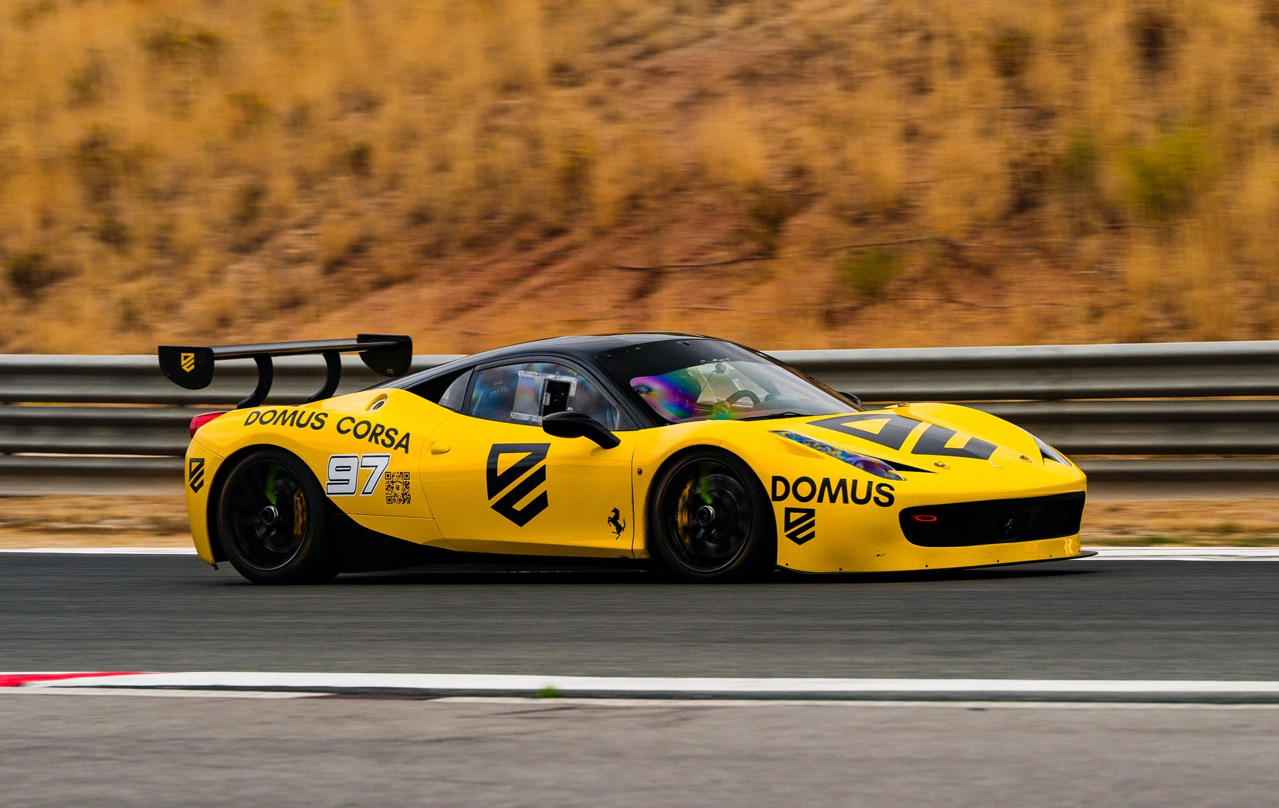 Yellow race car with the number 97 and sponsor logos driving on a race track, with a blurred background of dry grass and hills.