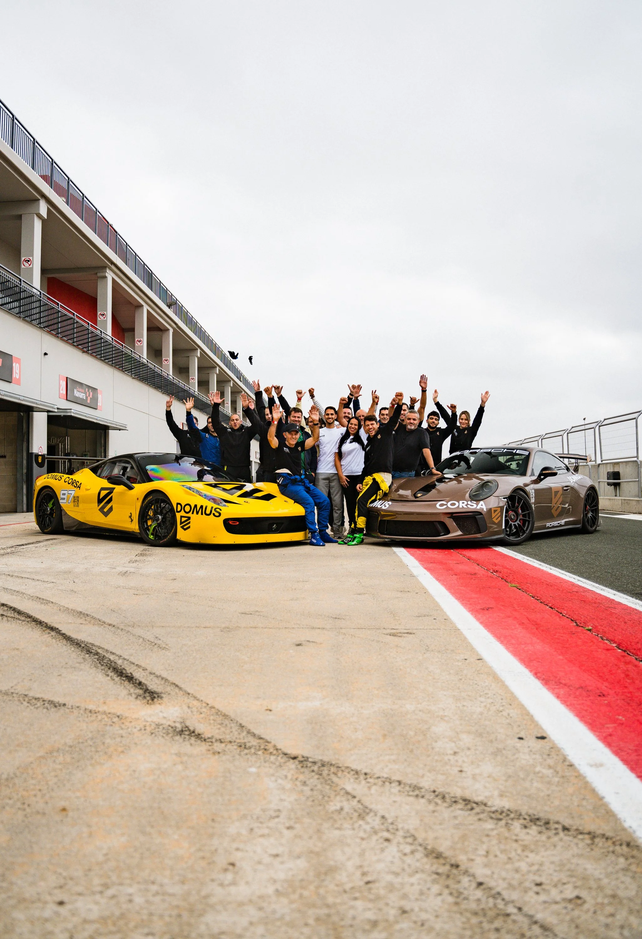 A group of people standing on a race track, celebrating with their hands raised, with two race cars parked in front of them and a building with balconies in the background.