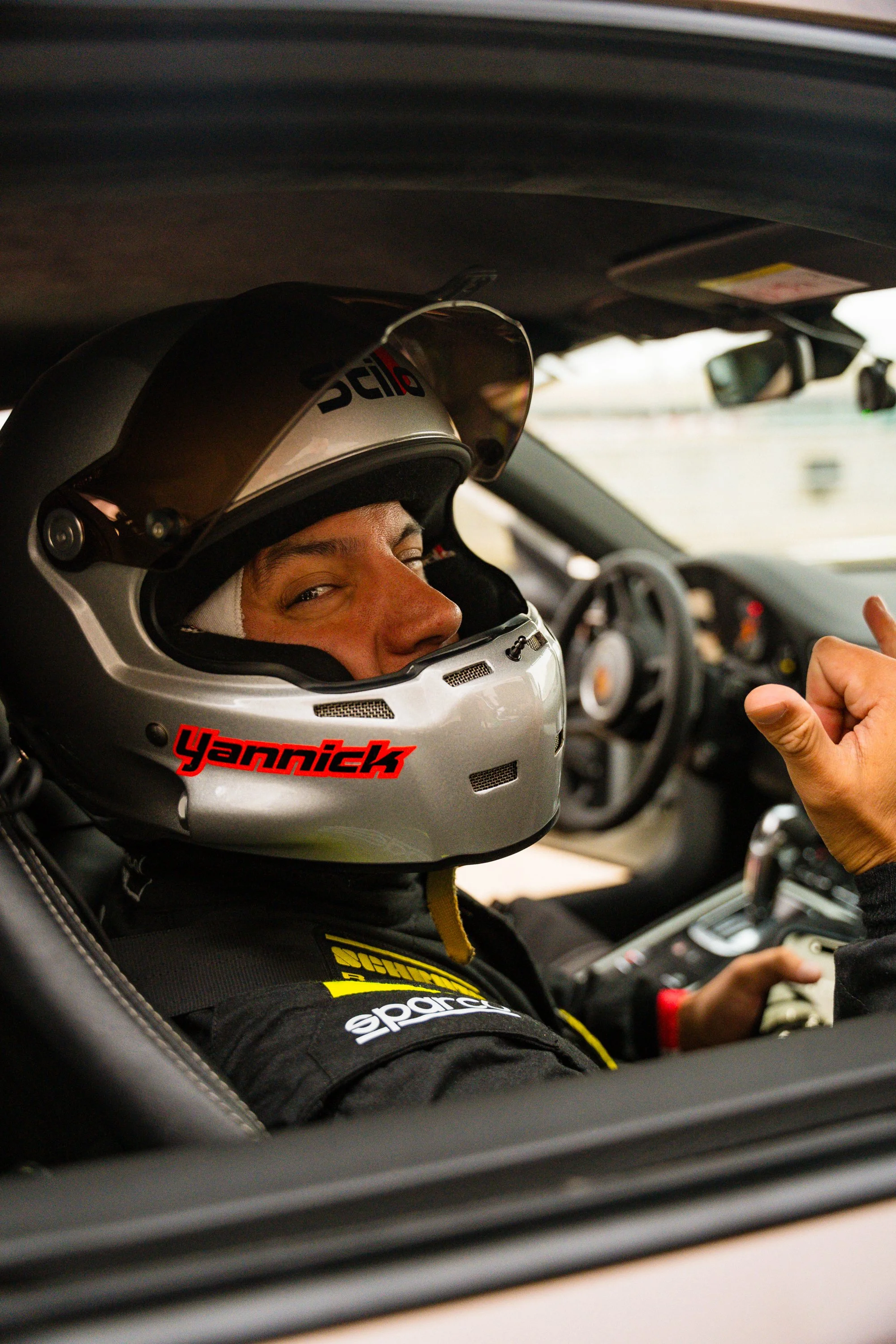 A race car driver wearing a racing helmet and suit sitting inside a car, giving a thumbs-up gesture.