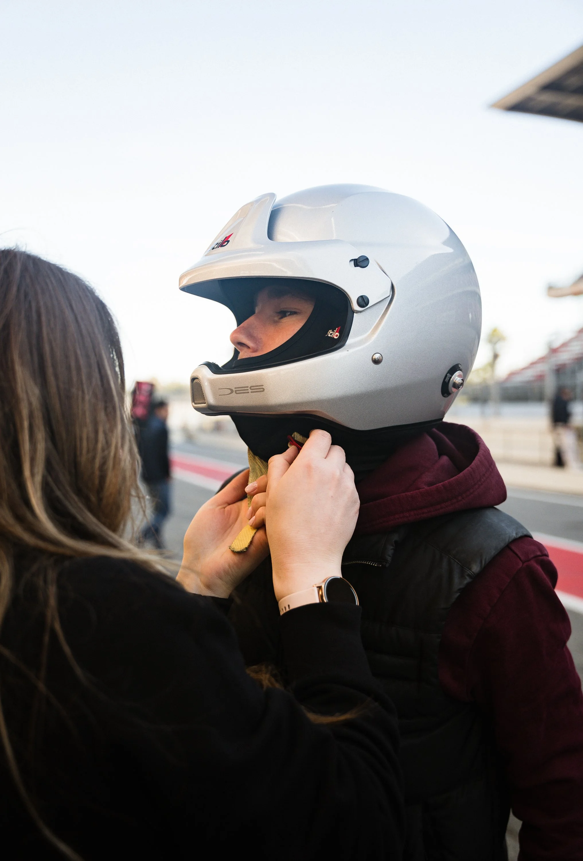 A person helps another person put on a silver motorcycle helmet with a black interior.