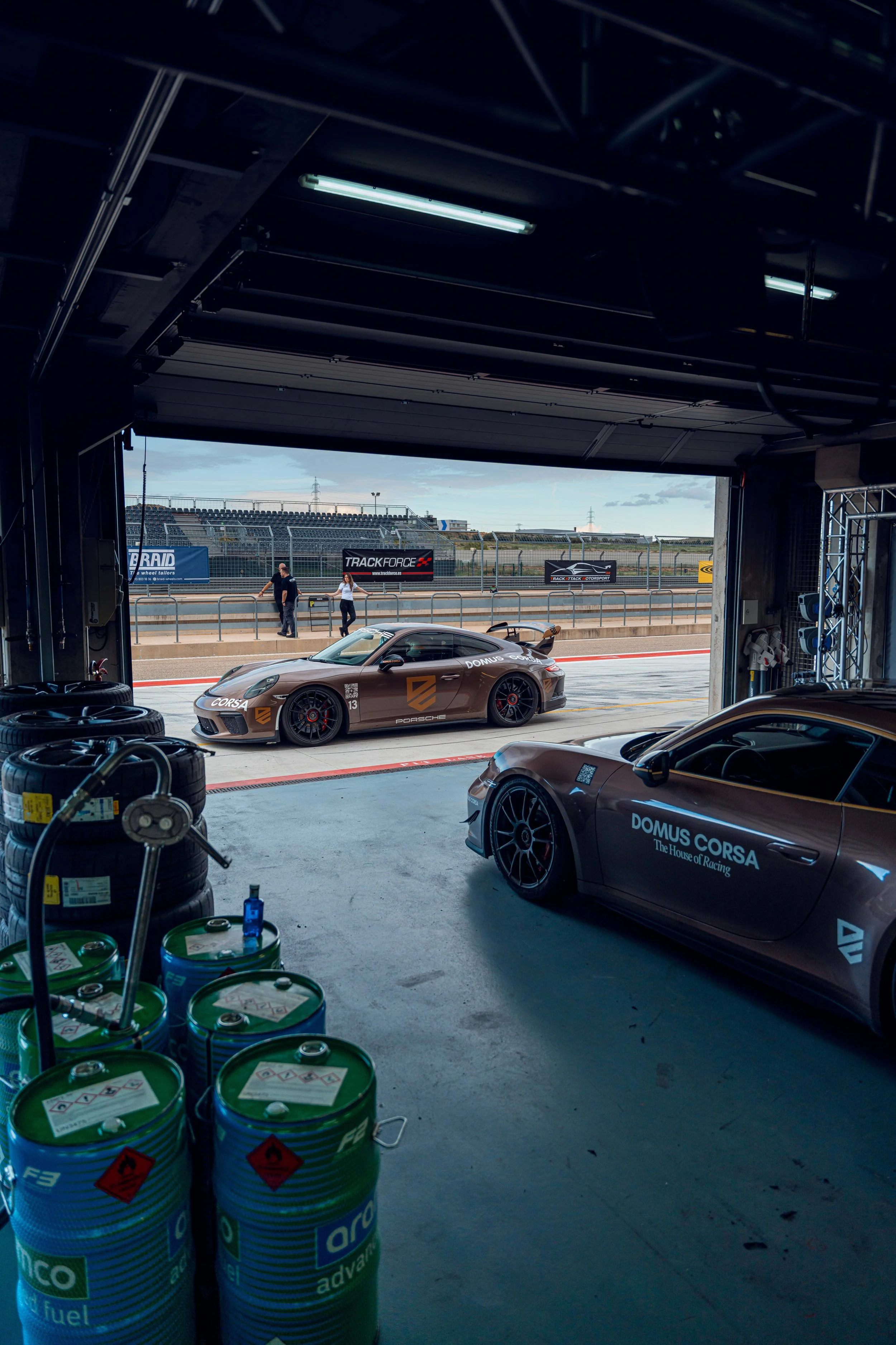 View from inside a garage or pit lane at a race track, with racing cars and tires visible. Outside the garage, a race car with the words 'DOMUS CORSA' on the side is parked, and two people are walking on the track. There are banners and fences in the