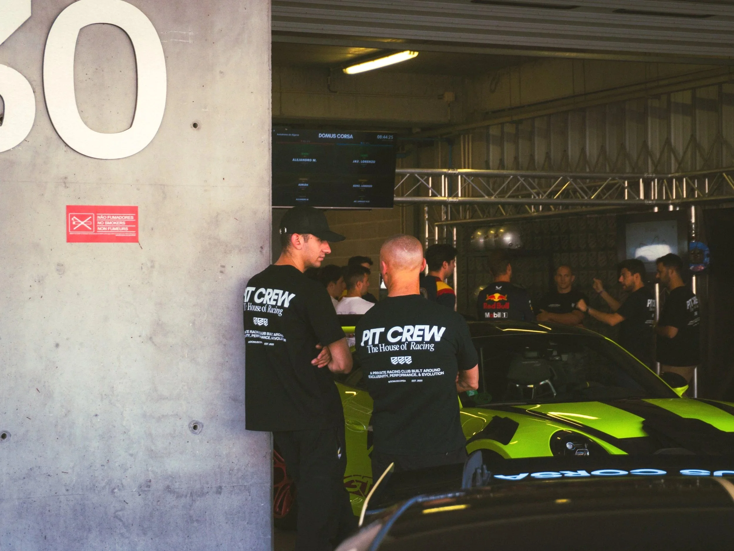People gathered around bright yellow sports cars with racing decals at a racing event. Some wear shirts labeled 'Pit Crew' and other racing gear. A digital screen displays race details in the background.