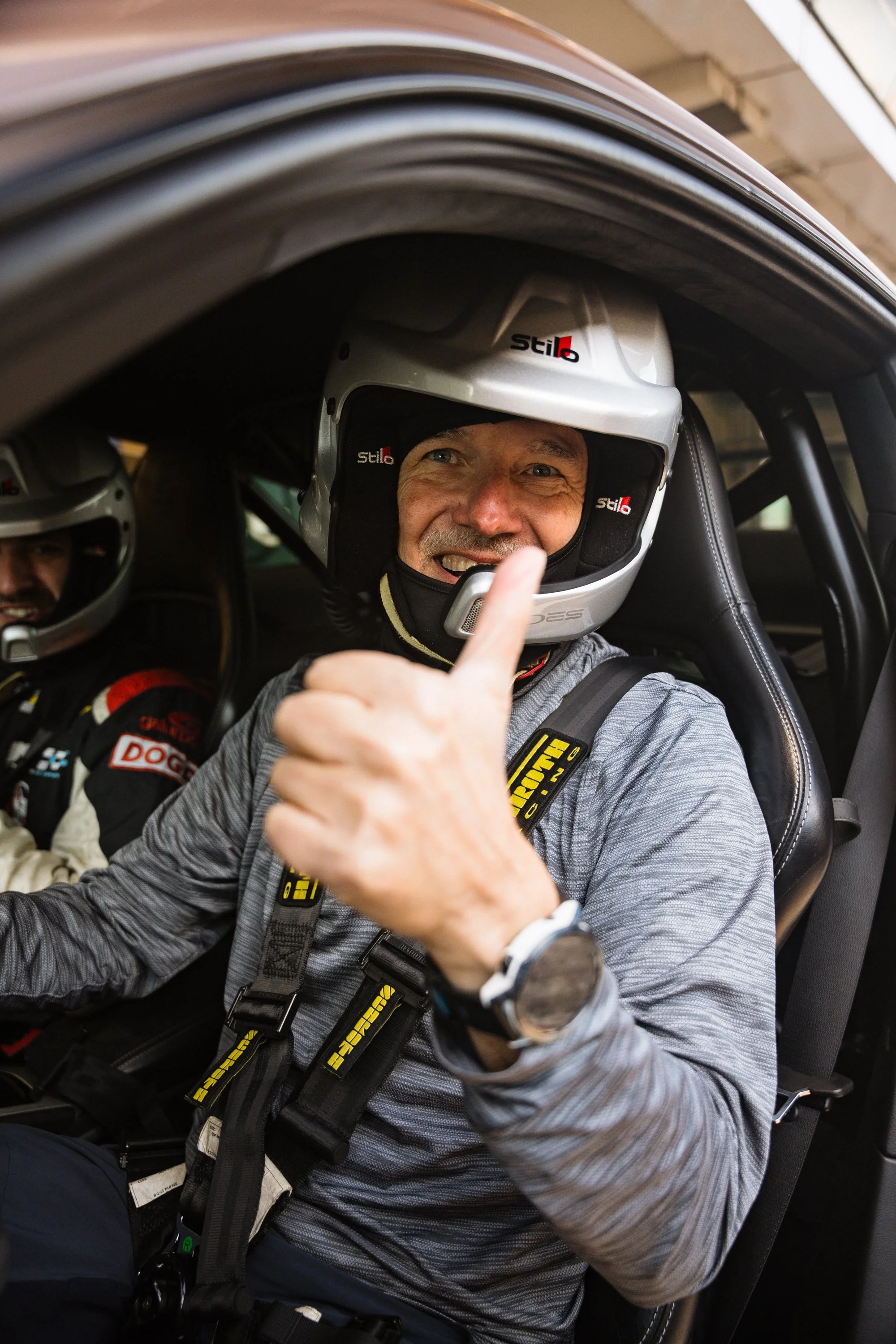 Two men inside a race car, wearing white helmets with the 'stilo' logo, smiling and giving a thumbs-up.