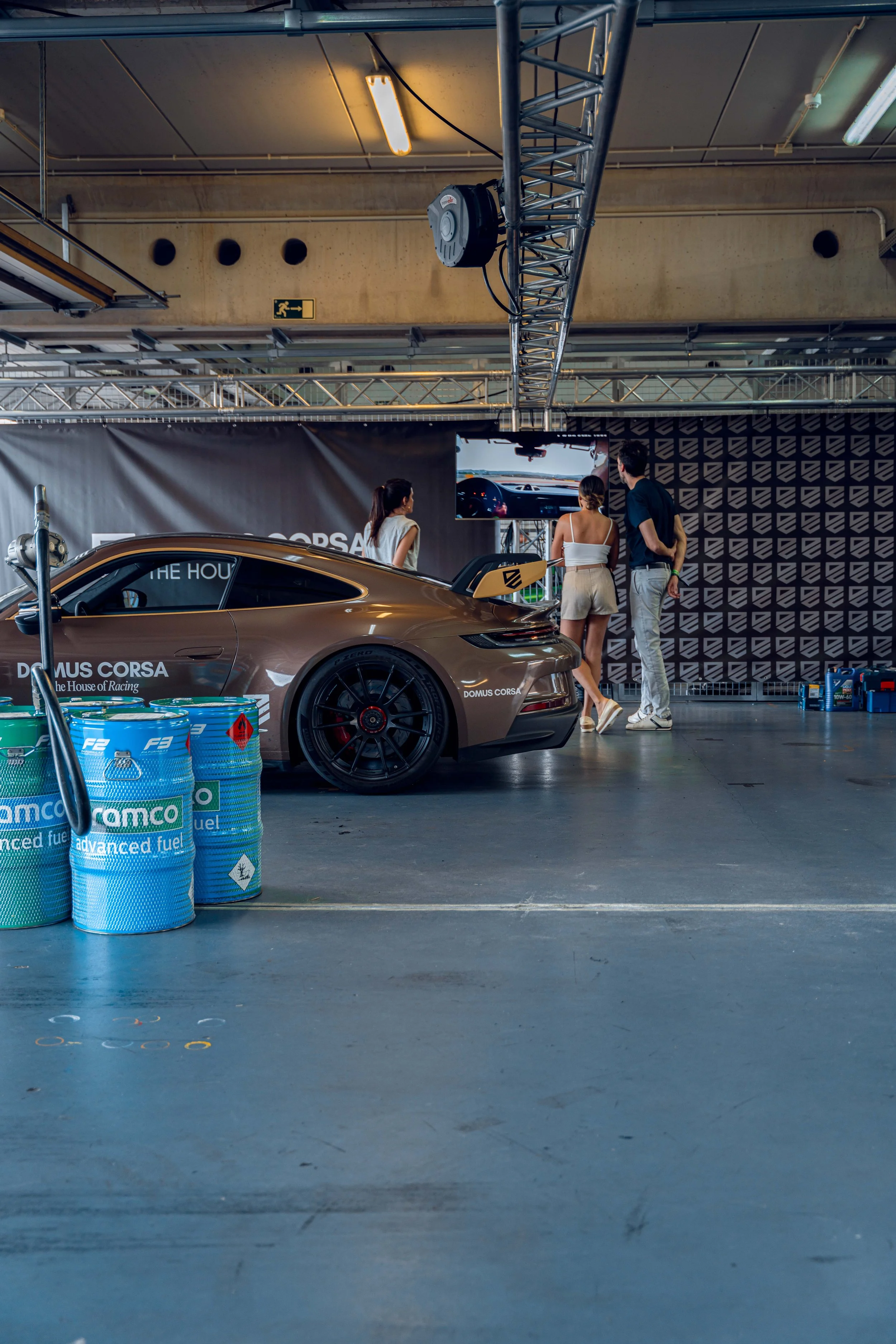 Three people standing in front of a racing car at an indoor event with fuel barrels nearby. The car has a rear wing and doors with branding. A large screen displays driving footage. The ceiling has exposed pipes and lighting fixtures.