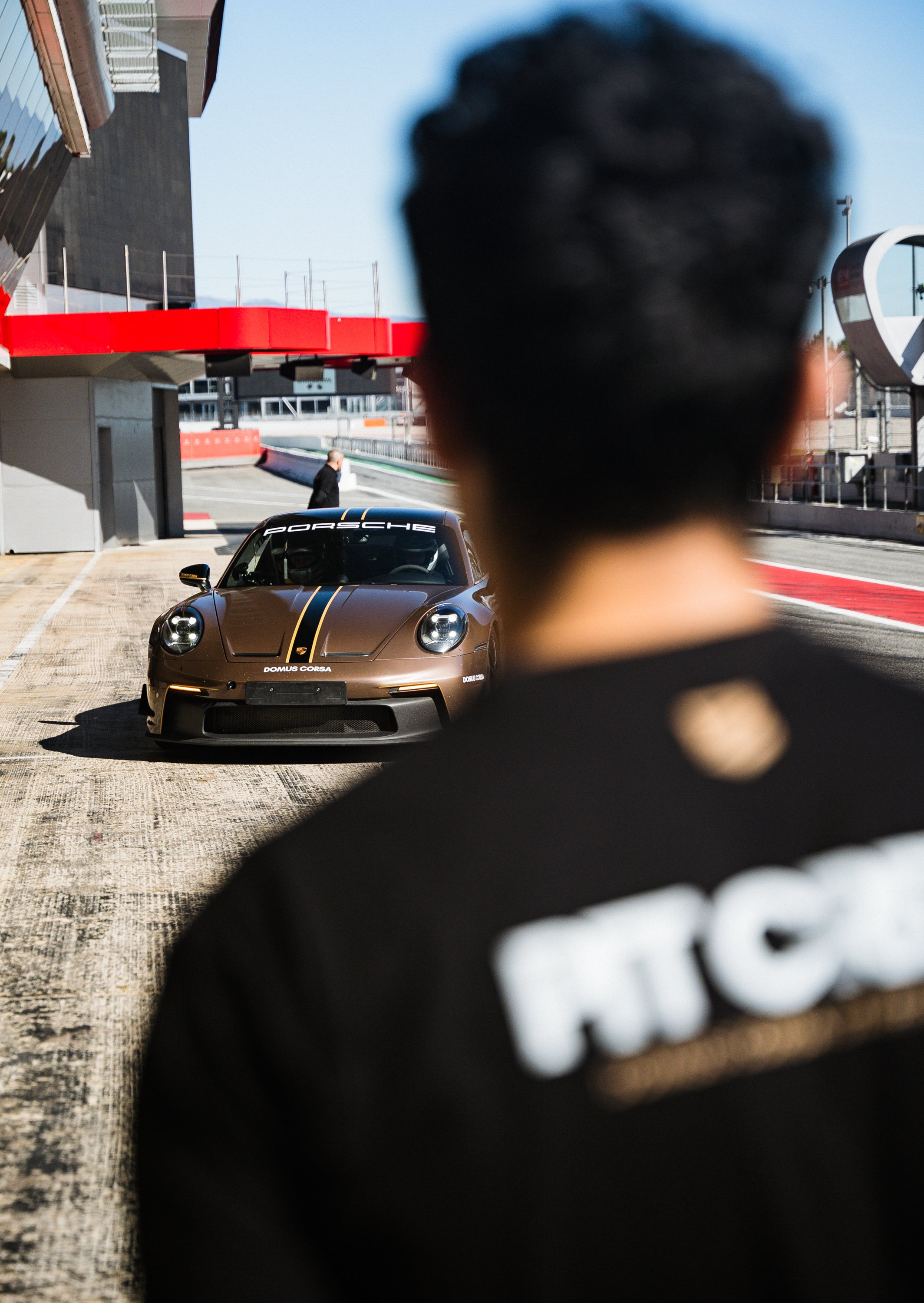 A man walking in front of a Porsche race car on a racetrack, with the back of a person's head and shoulder in the foreground.