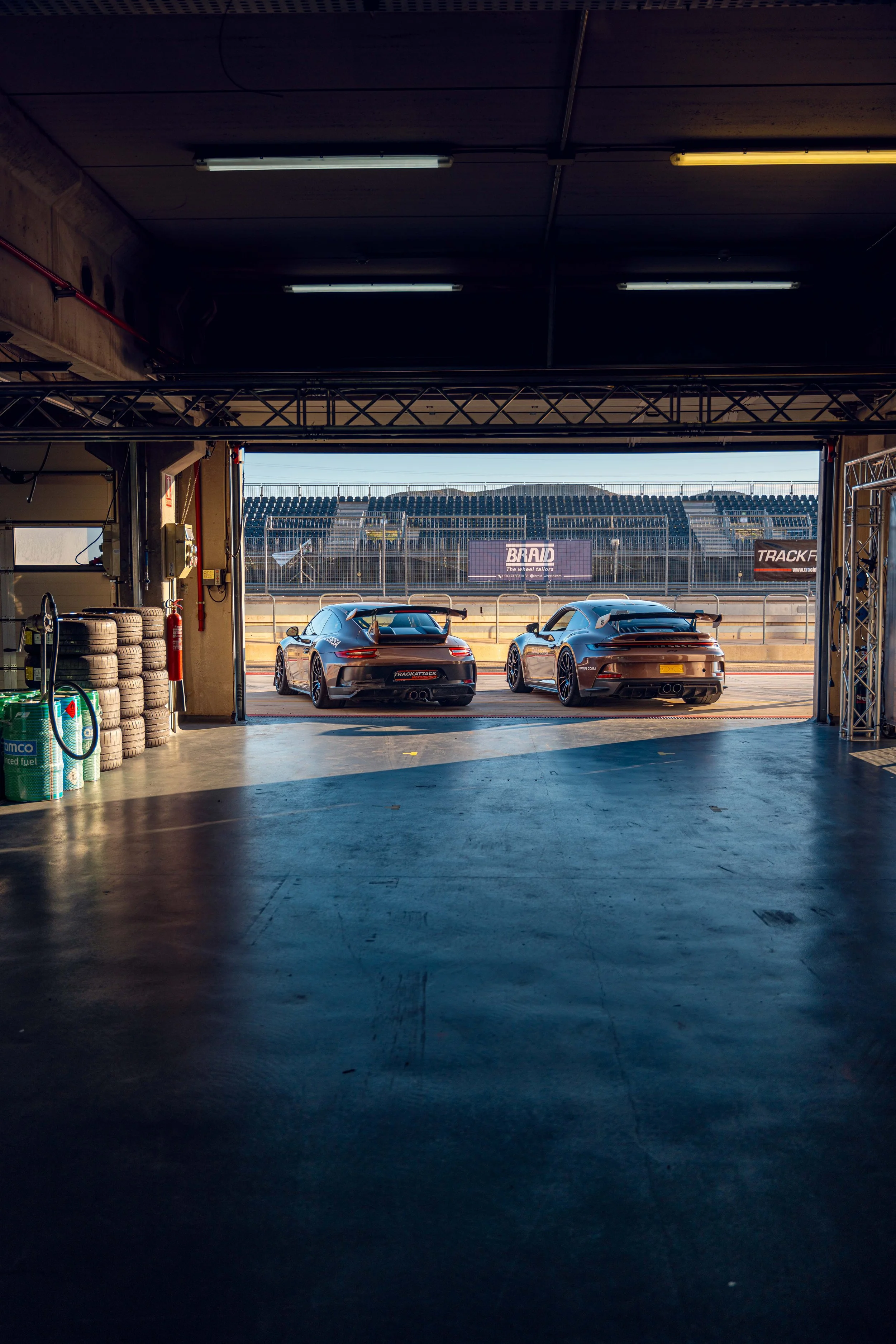 Race car garage with two black Porsche race cars parked outside, tire stacks on the left, and grandstands in the background.