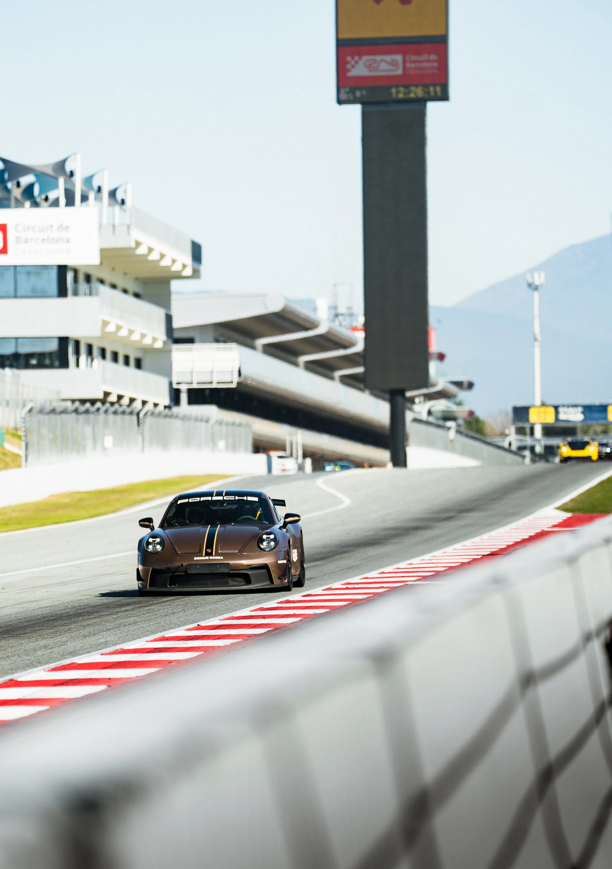 A brown Porsche race car on a racetrack with red and white curbs, under a blue sky, with a modern stadium and mountainous background.