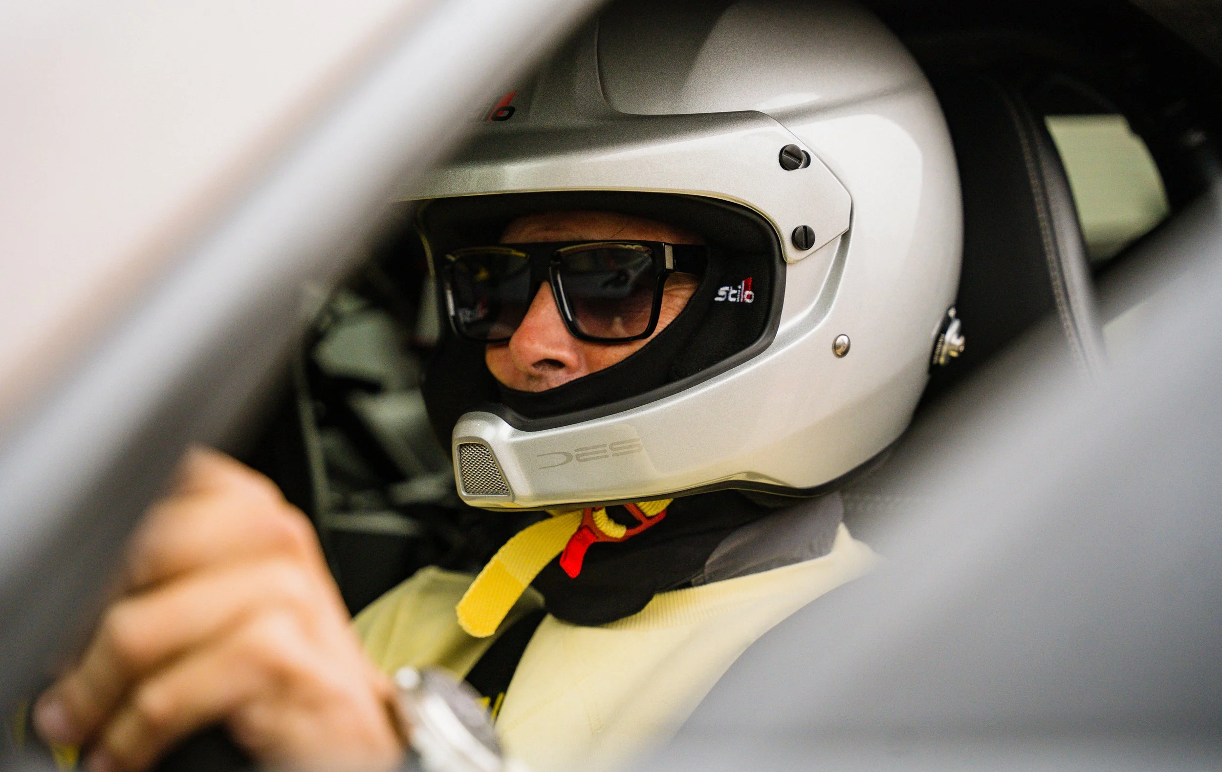 Close-up of a person wearing a racing helmet and sunglasses, seated inside a race car, with a focus on their face and helmet.