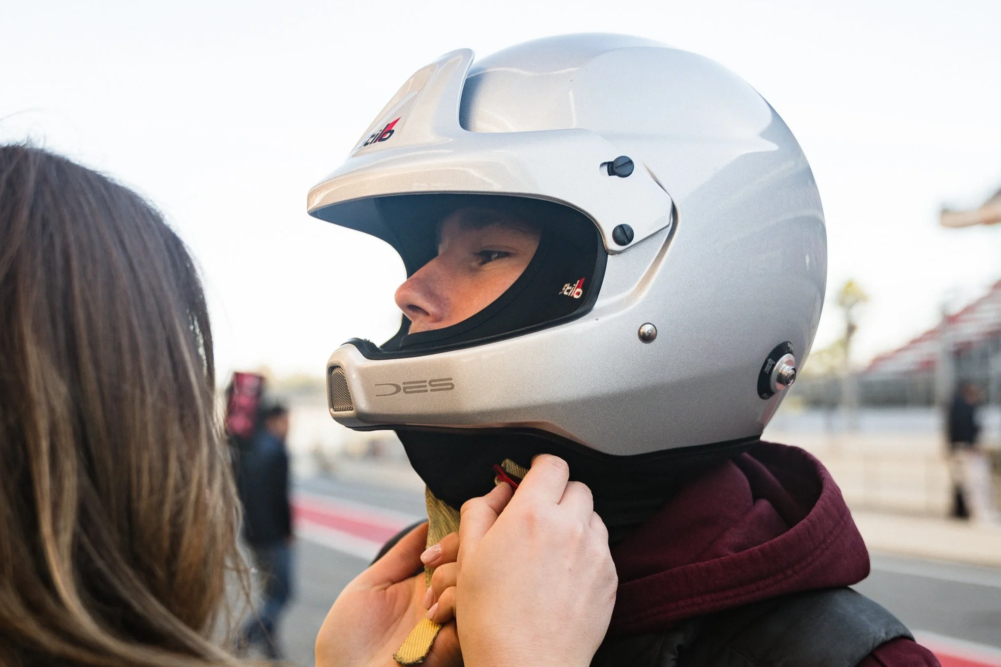 Person wearing a white motorcycle helmet, seen from the side, holding the chin strap while another person helps secure it.