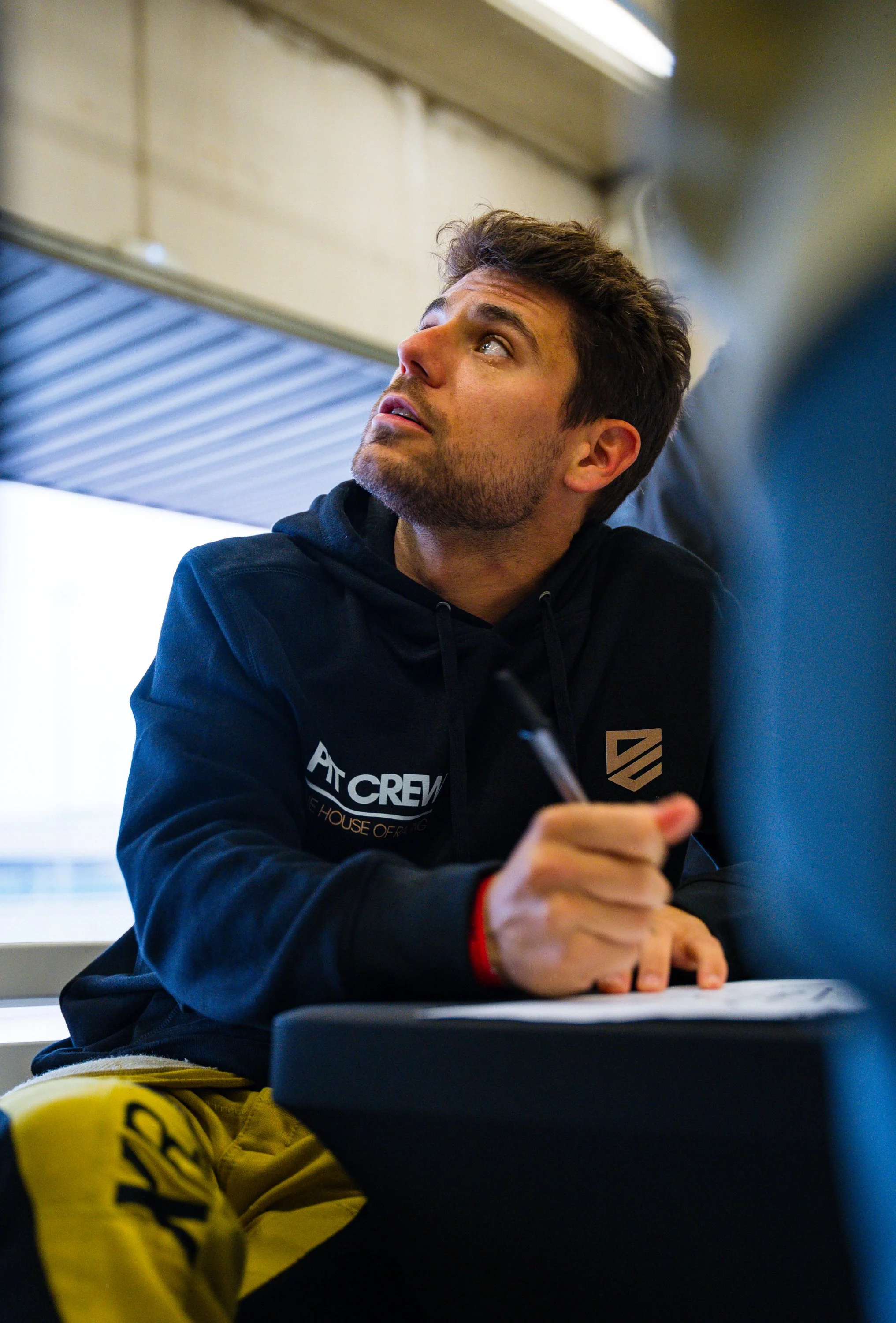 A young man with brown hair and a beard, wearing a black hoodie with sponsor logos, sitting at a table in a room with a window and roof overhang. He is looking to his left, with a pen in his right hand and papers in front of him.
