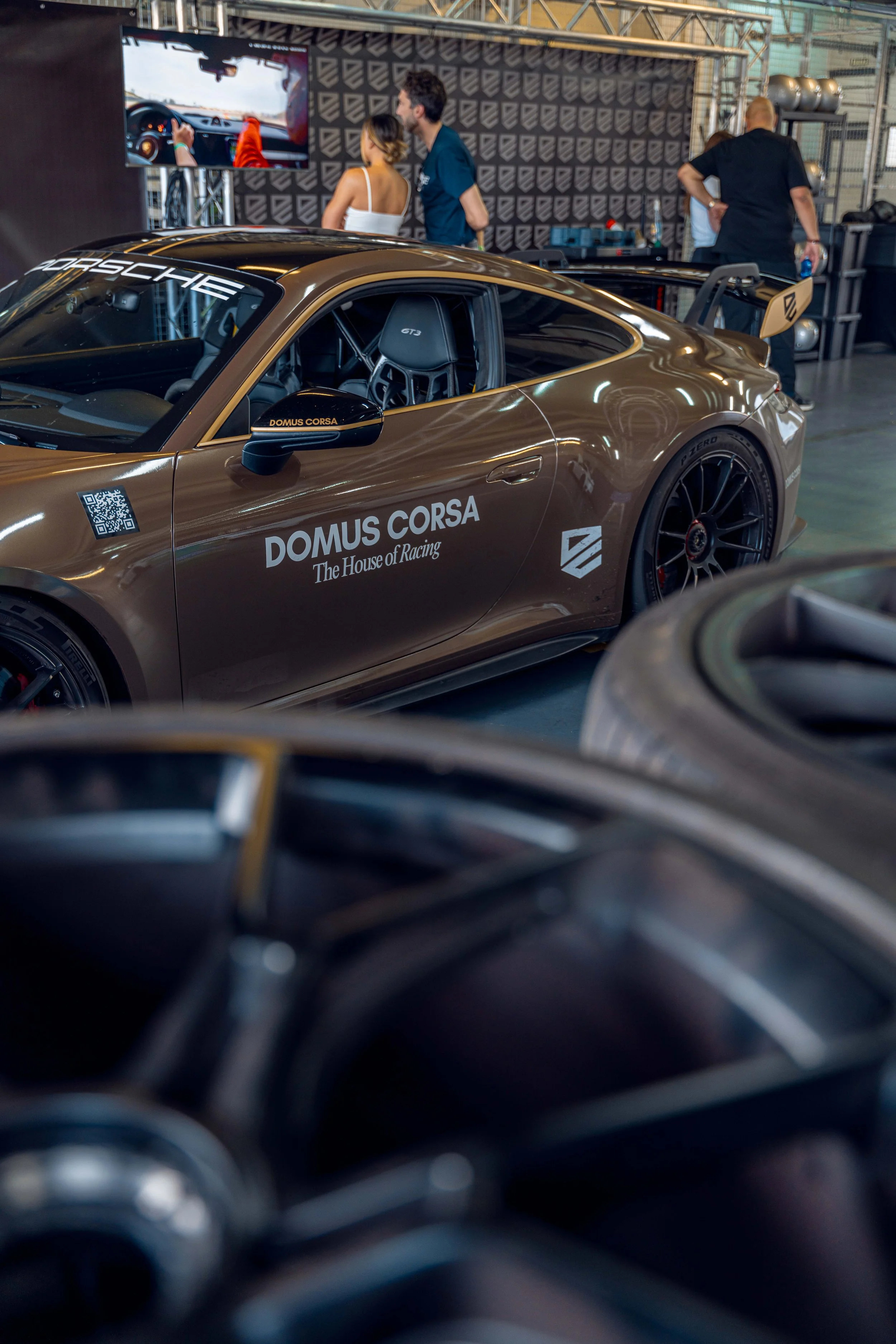 A brown Porsche race car with racing decals, displayed indoors at an event, with several people in the background viewing a monitor and talking.
