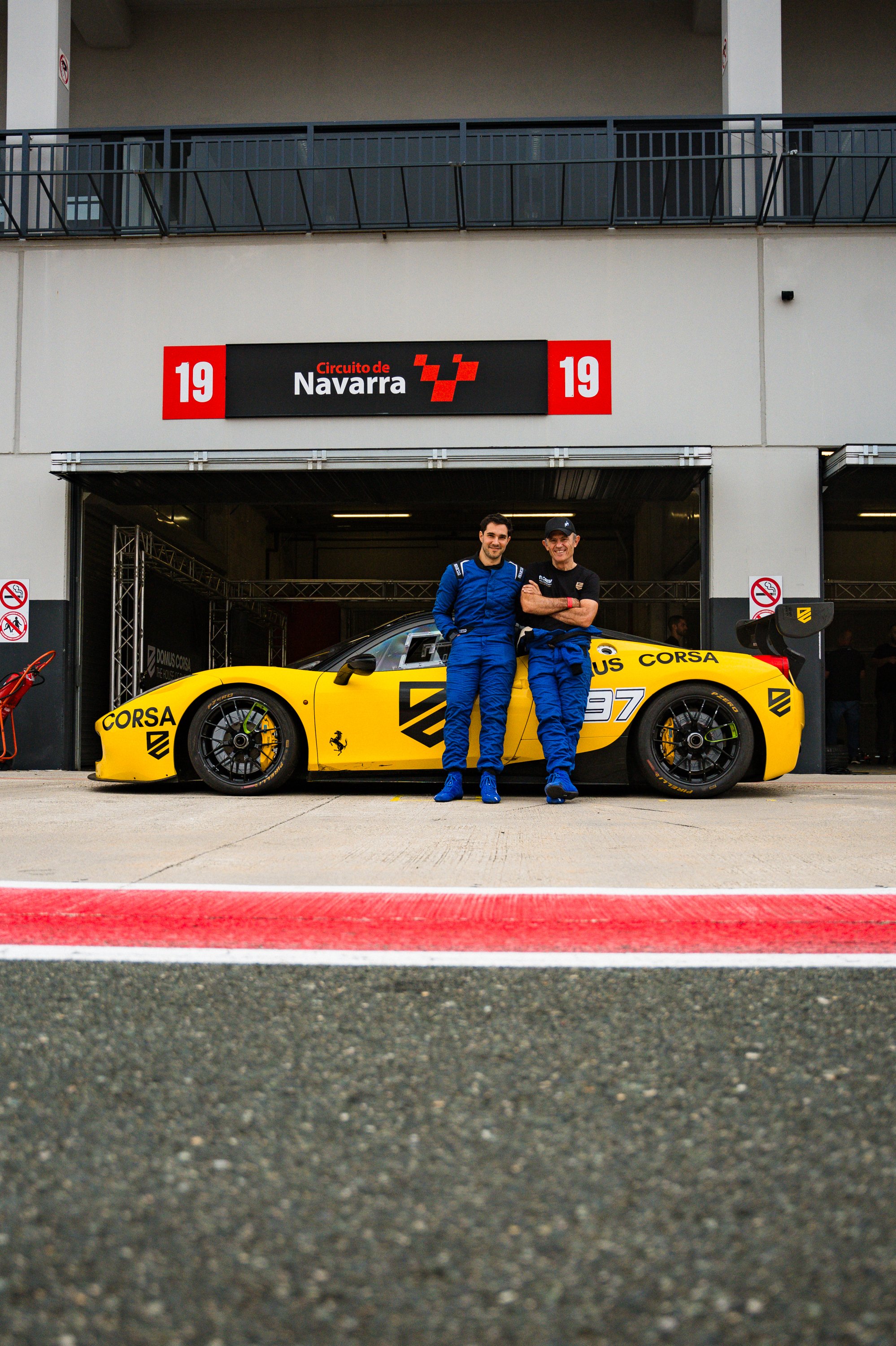 Two men in racing suits standing next to a yellow race car with sponsorship branding, inside a garage at a racing circuit with a sign reading 'Circuito de Navarra' above.