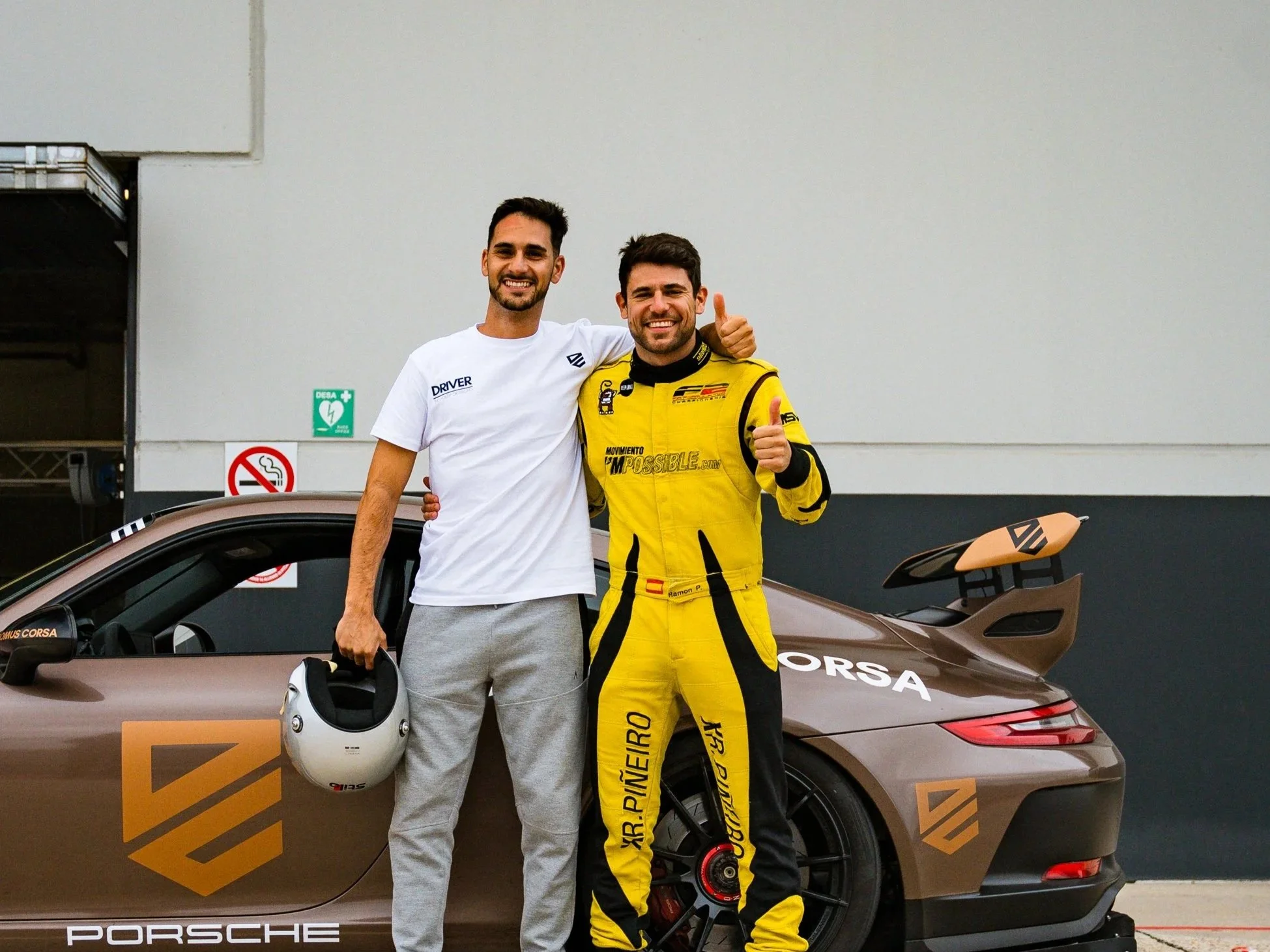 Two men standing together beside a racing car, one in a white shirt holding a helmet, the other in a yellow racing suit giving a thumbs-up, smiling.