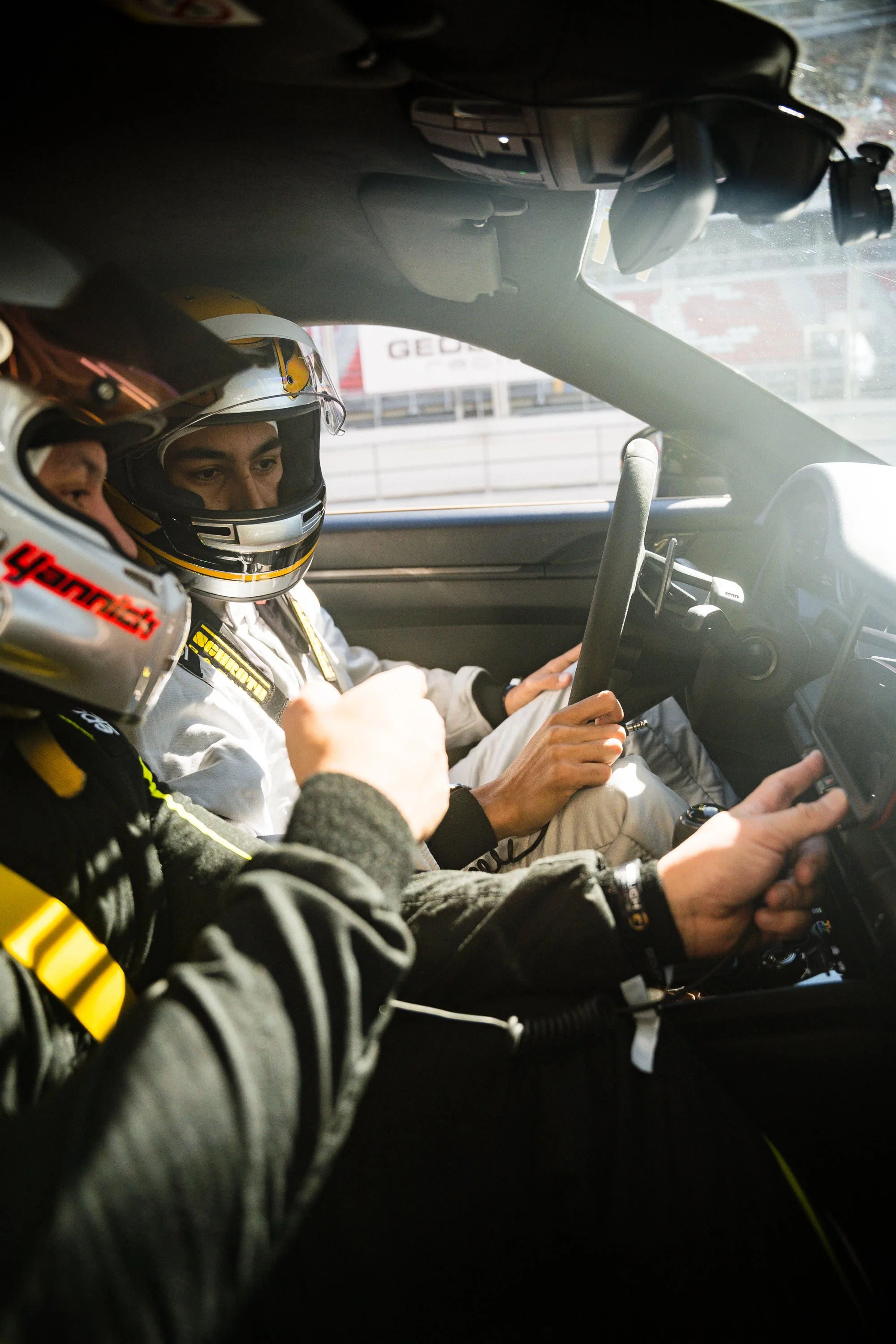 Inside a race car with two drivers wearing helmets and racing suits, focused on the dashboard and steering wheel.