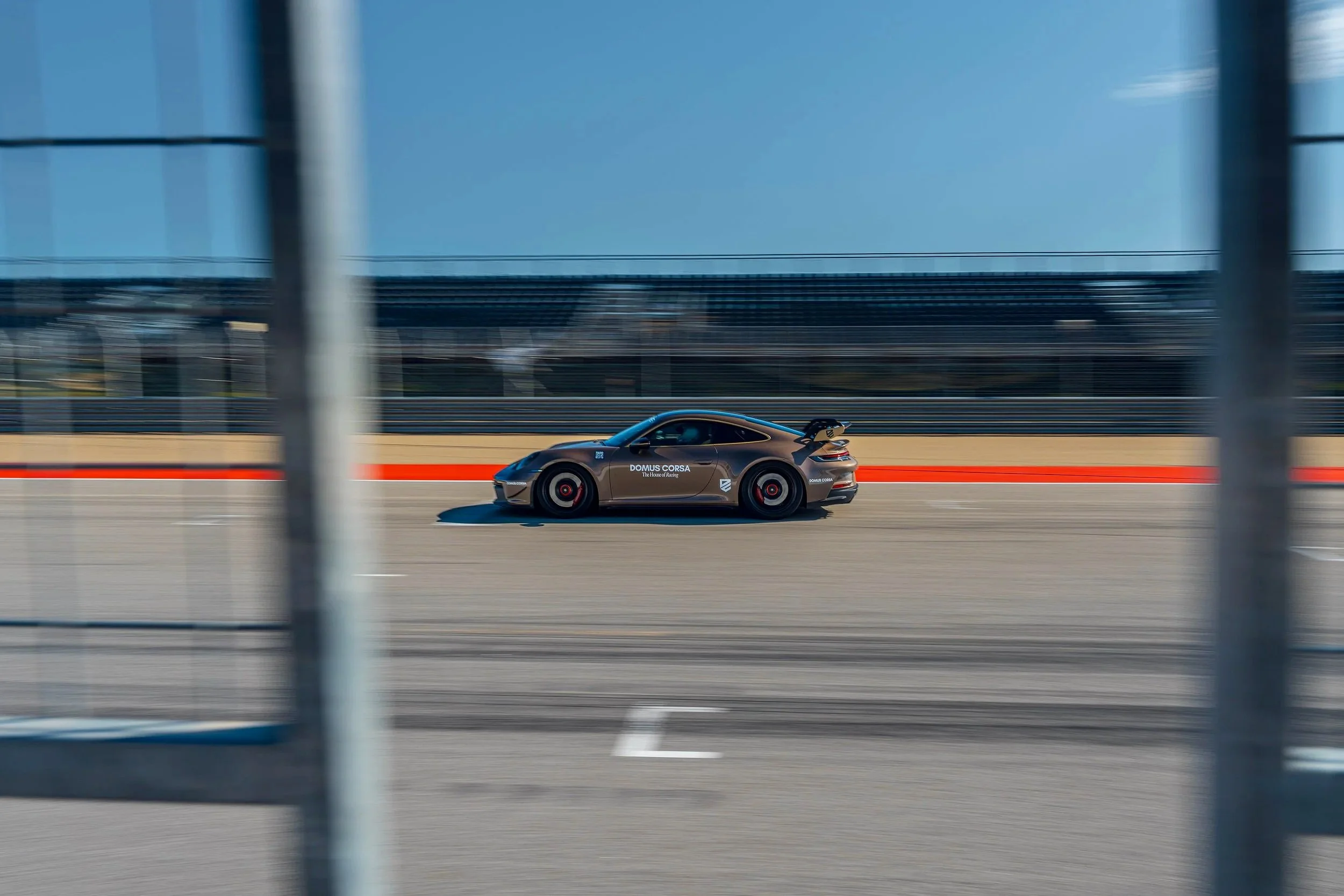 A dark gray sports car racing on a track, viewed through metal fencing posts with a blurred background of a blue sky and stadium stands.