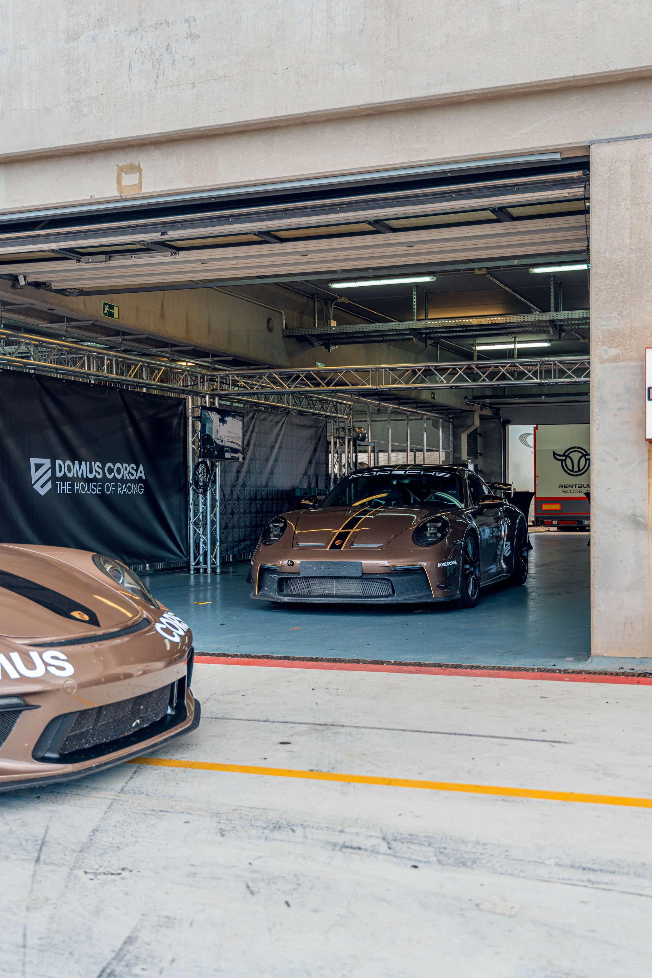 A black Porsche race car parked inside a garage area with a black banner that reads 'Domus Corsa the house of racing' and a poster showing a racing vehicle. The garage has open sides and is surrounded by a concrete structure.