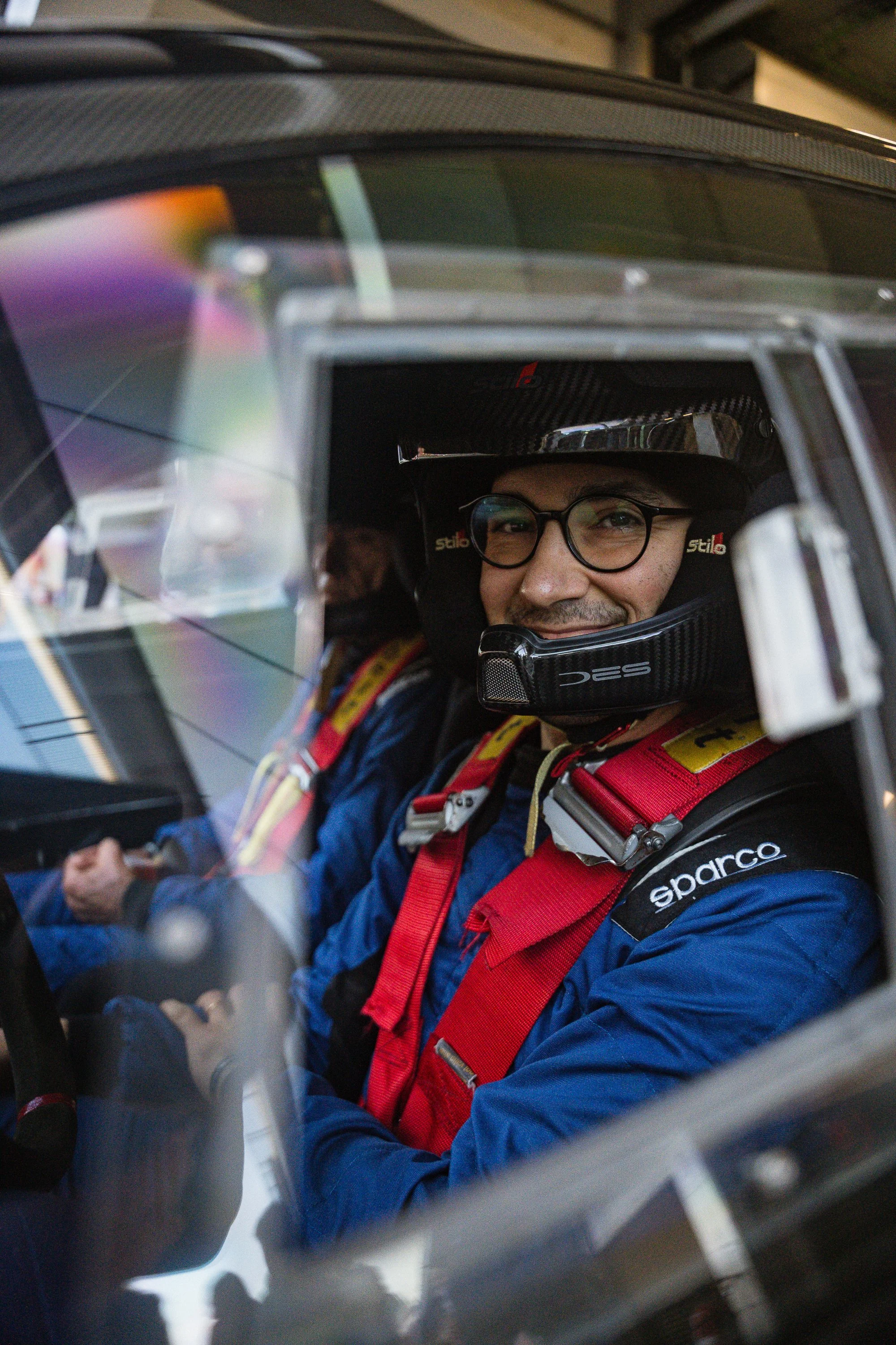 A man wearing glasses and racing gear, including a Sparco helmet and a racing suit, sitting inside a race car, smiling and looking out of the window.