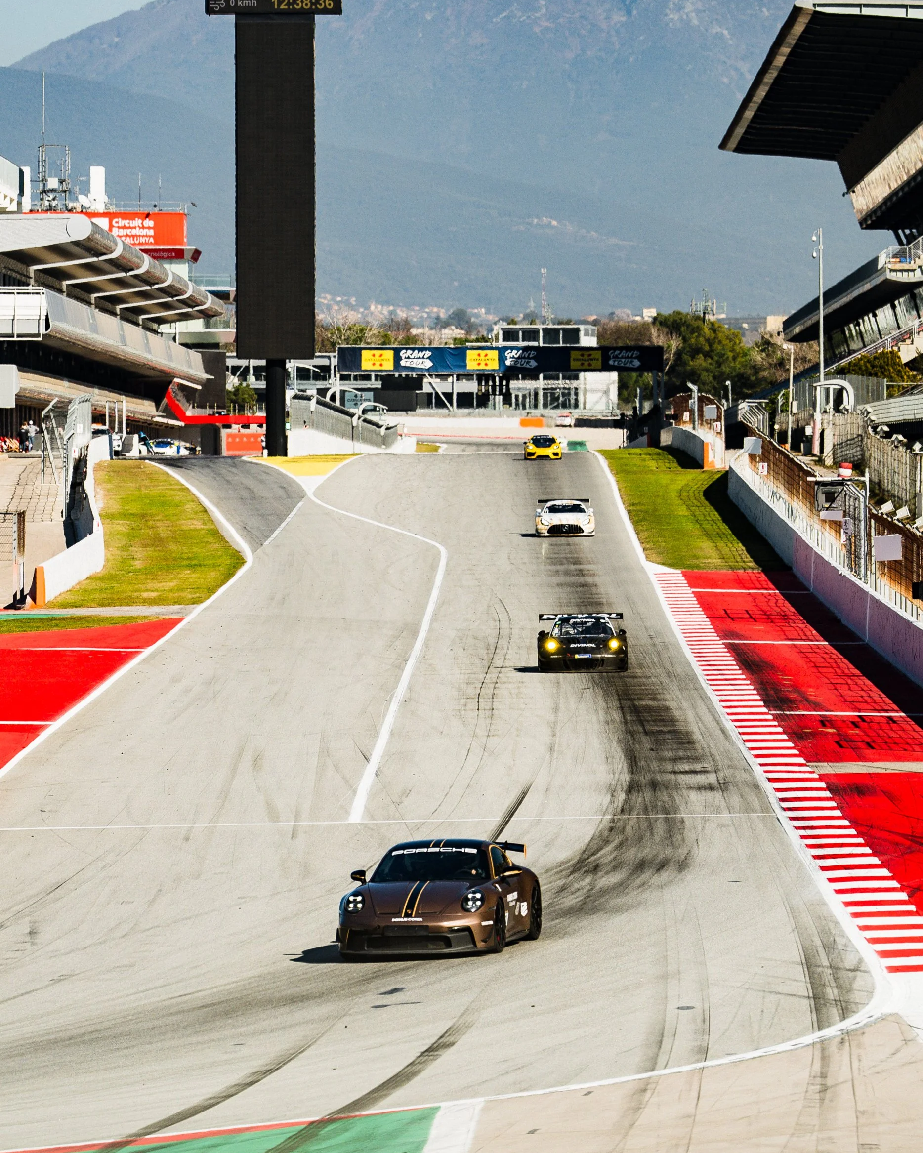 Multiple race cars driving on a track at the Circuit de Barcelona-Catalunya, with a mountainous background and digital display showing the time and speed.