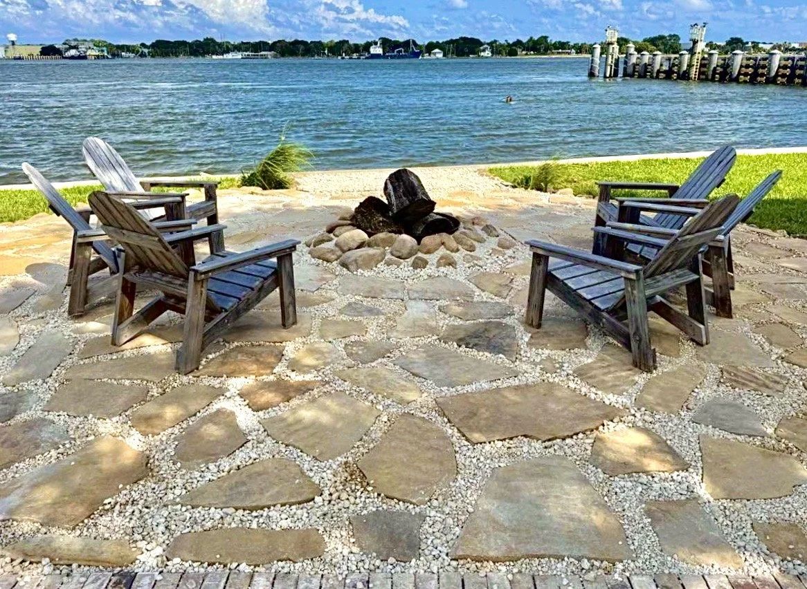 Four wooden chairs arranged in a semi-circle around a fire pit with large rocks and a spark screen, overlooking a body of water under a partly cloudy sky.