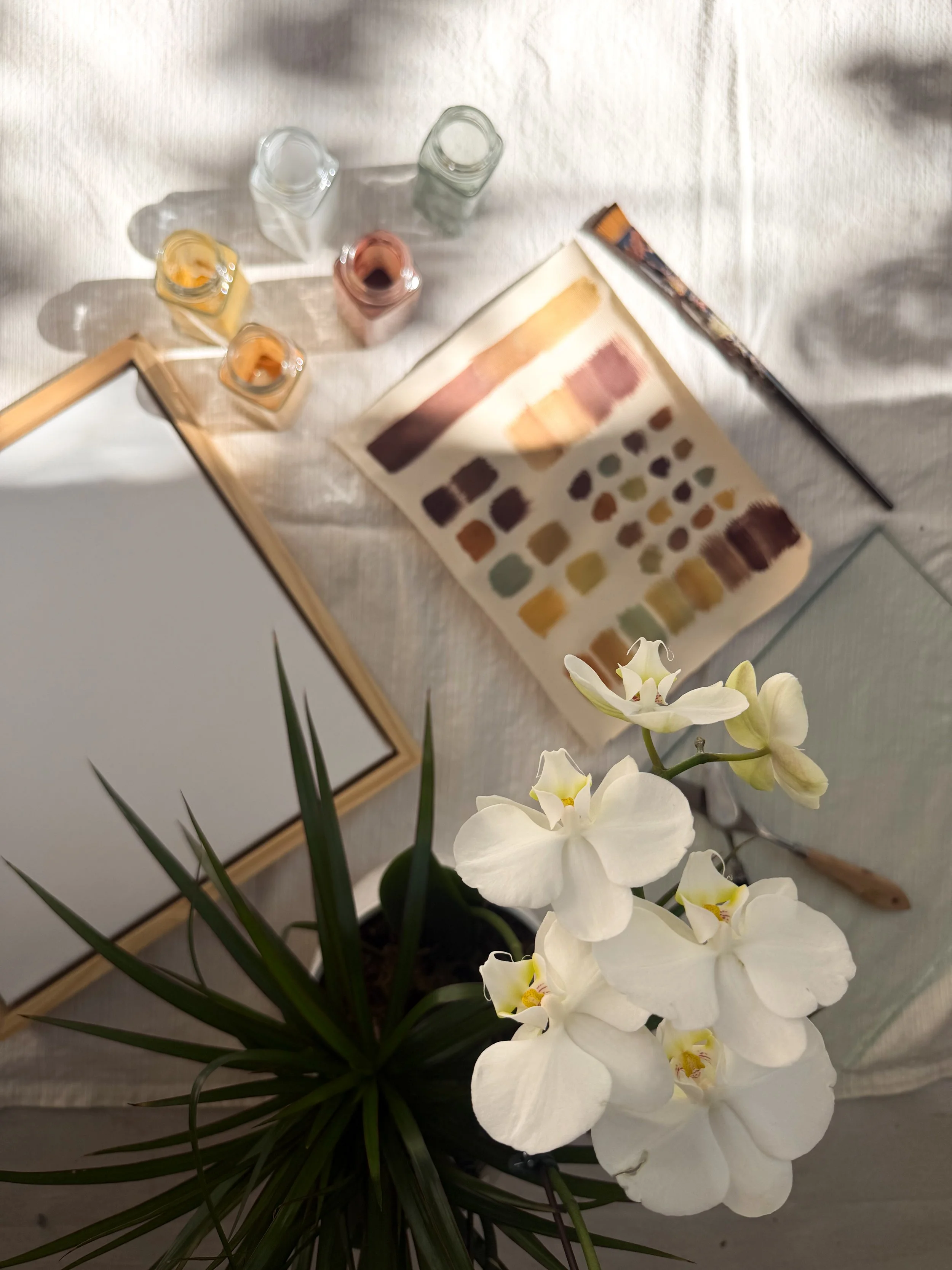 A bouquet of white orchids with green leaves in a white pot, placed on a light-colored surface on a cream tablecloth with a square, blank canvas framed in light wood, a color swatch, a paintbrush, and small glass jars of pigment nearby.