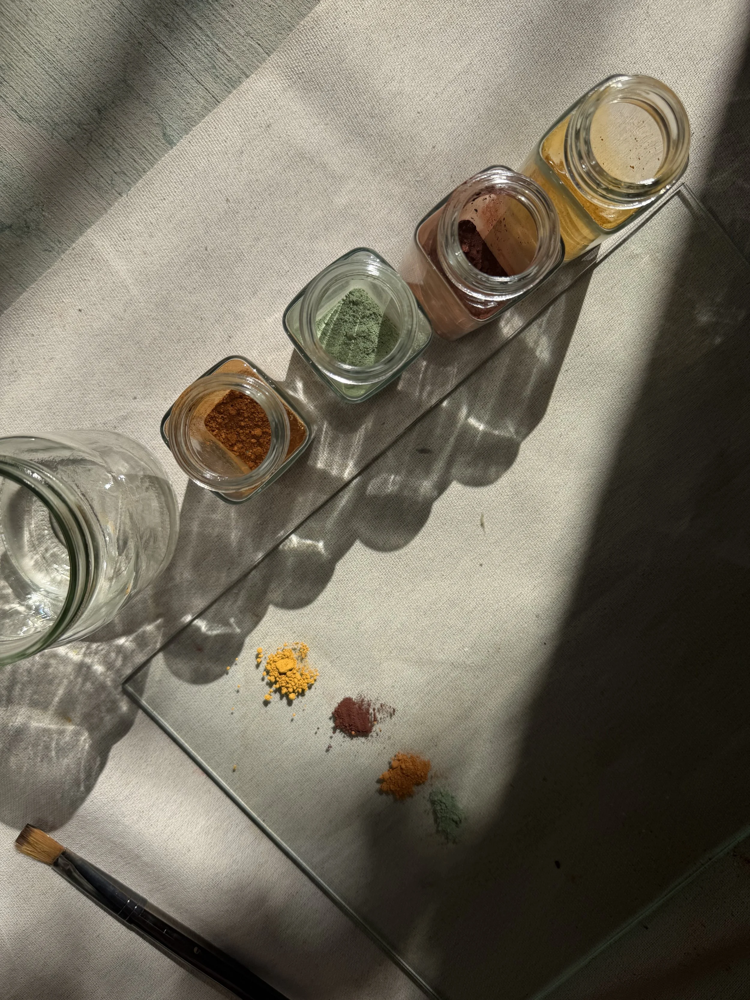 Small glass jars containing colorful pigments arranged next to a glass palette on a light-colored cloth surface, with some spilled pigments on the glass palette.