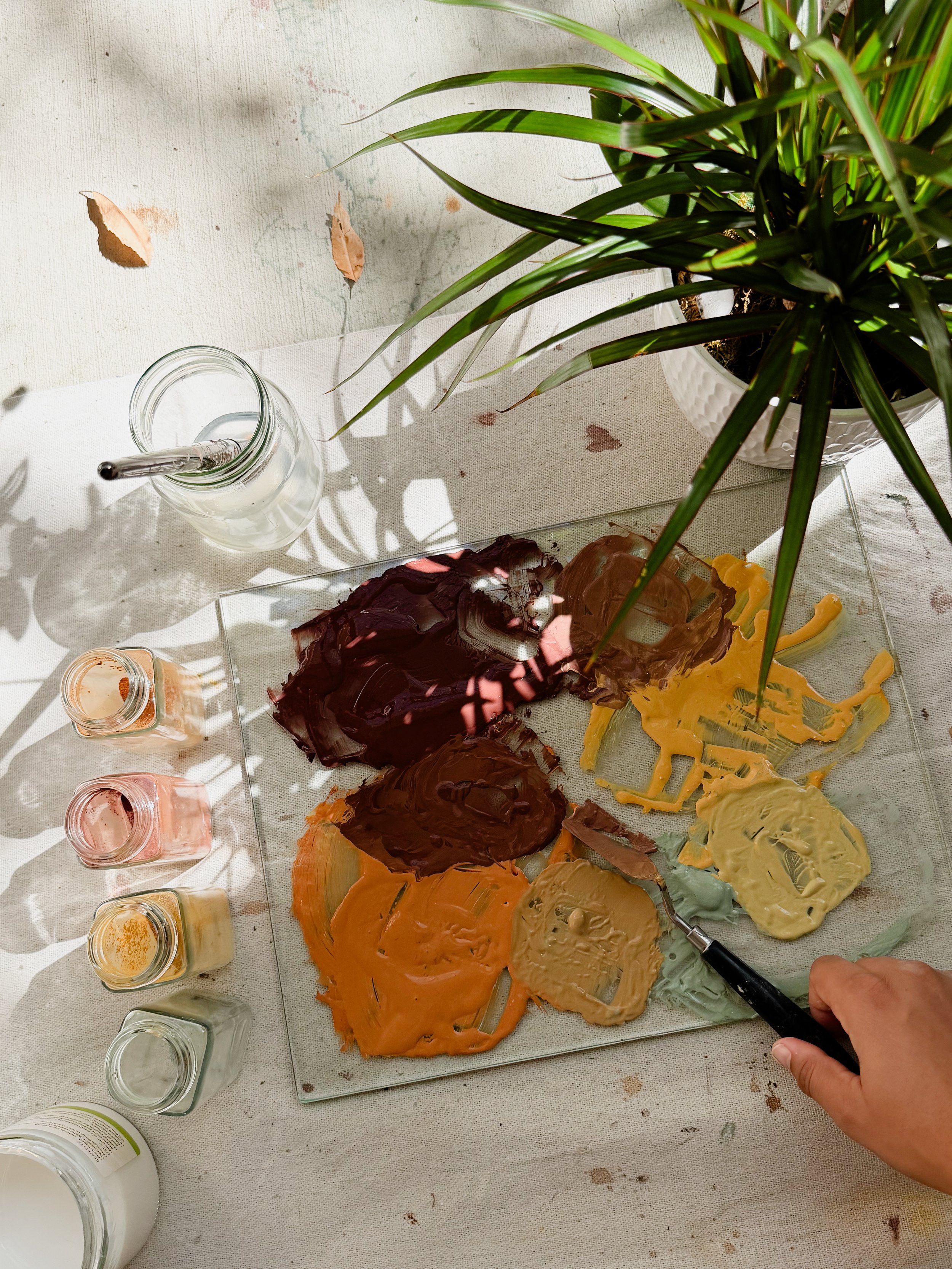 A person's hand holding a palette knife with yellow, beige, and brown paint, surrounded by various jars of pigment in colors like orange, green, and red, and a glass jar with a paintbrush, on a white surface with a large houseplant nearby.