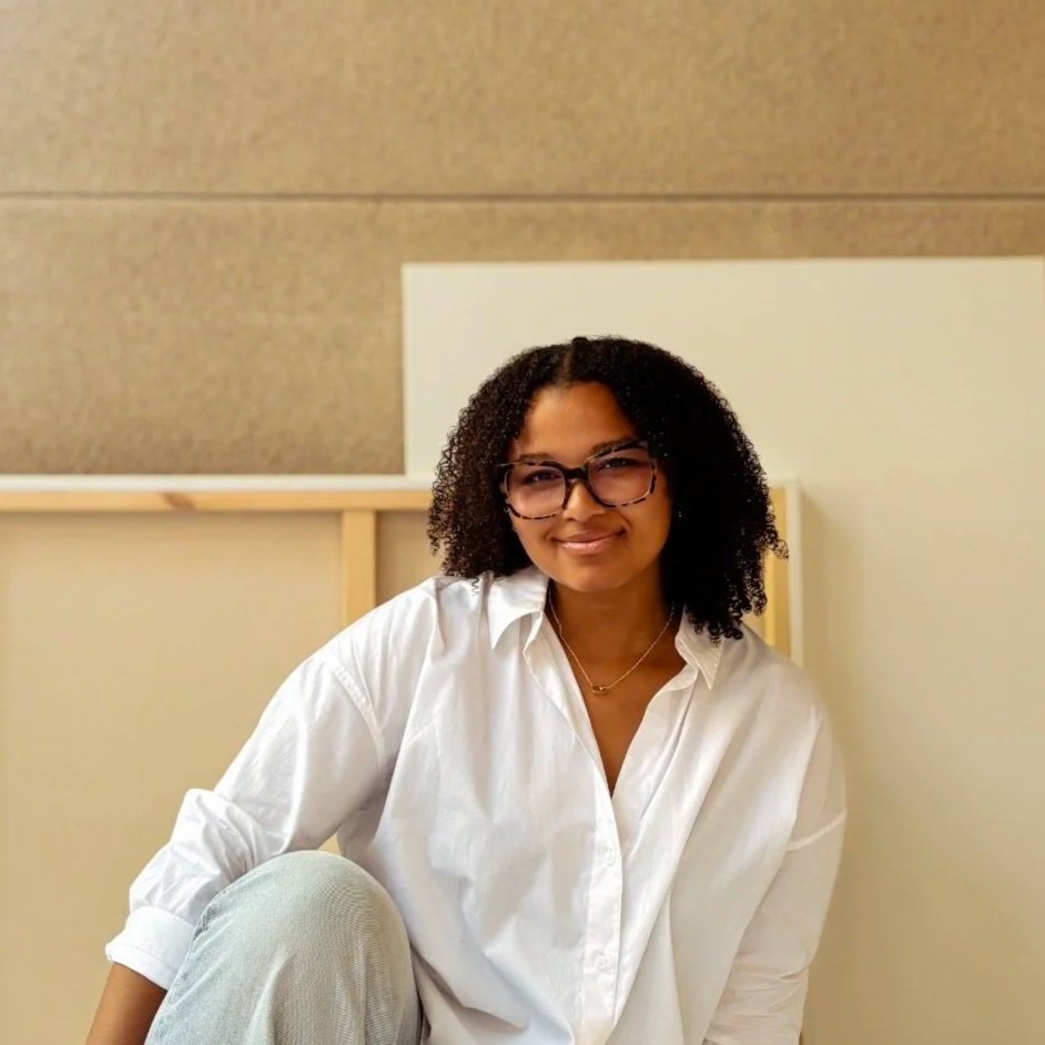 A woman with curly hair wearing glasses and a white button-up shirt, sitting in front of a beige background with a blank canvas behind her.