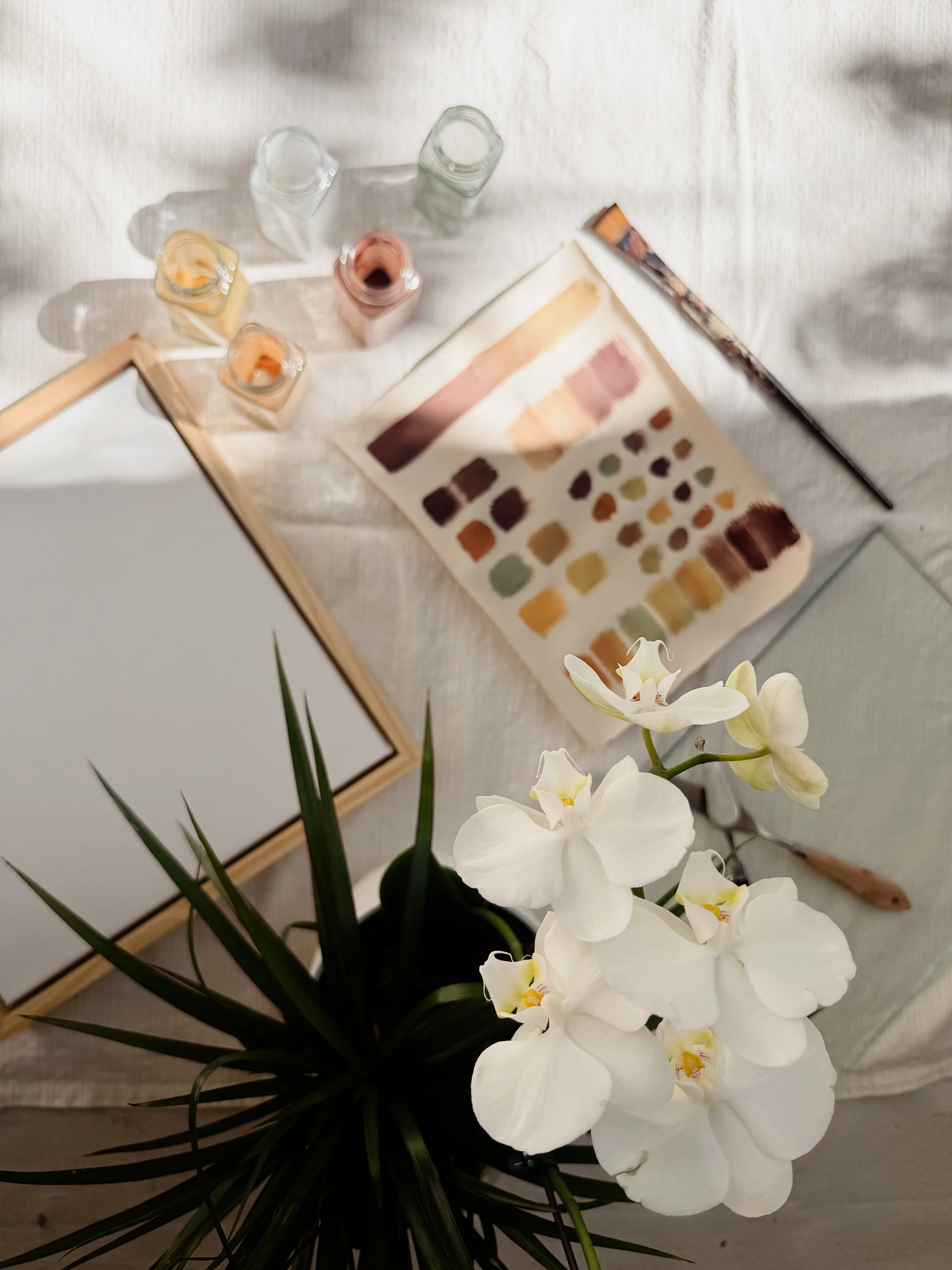 A bouquet of white orchids with green leaves placed on a table with art supplies including a paint palette, paintbrushes, and glass jars of pigment, along with a blank framed canvas.