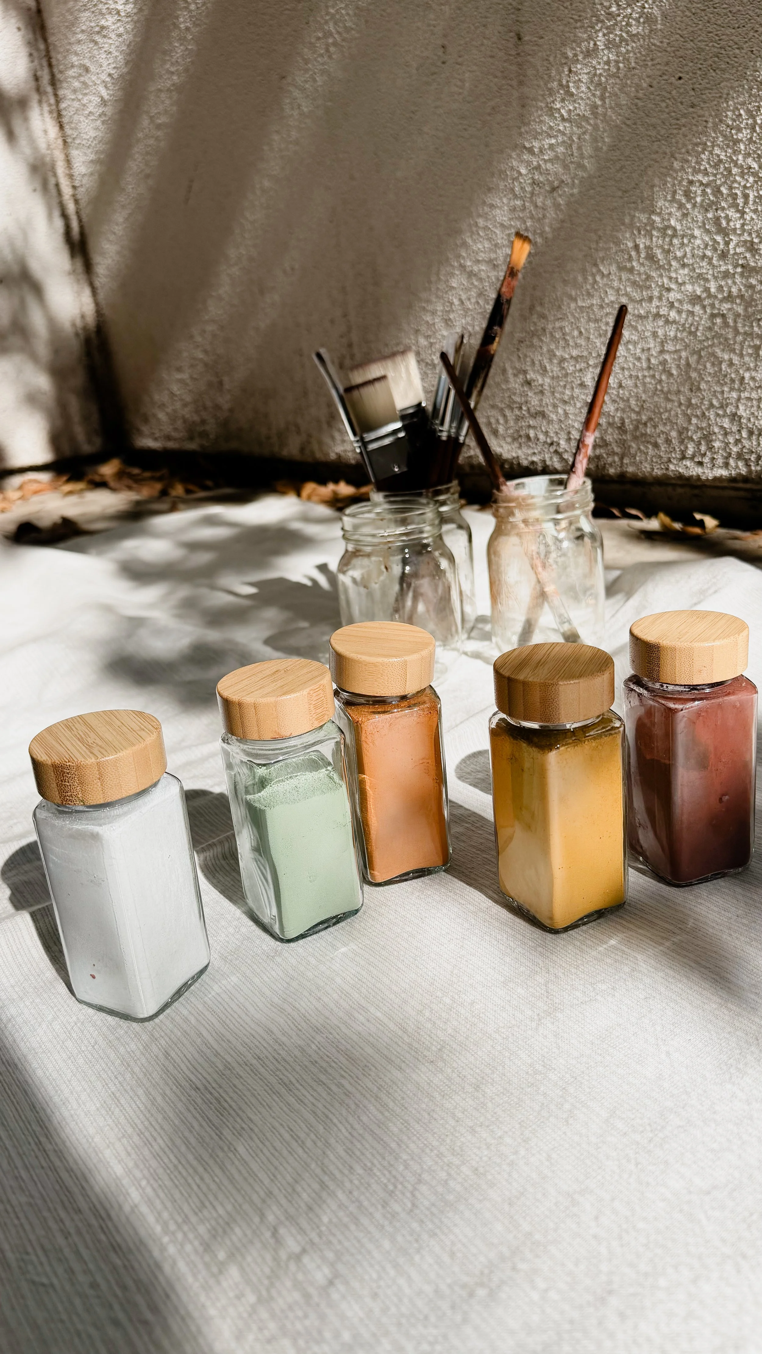 Glass jars with colorful powders and paintbrushes on a white fabric surface outside, sunlight casting shadows.