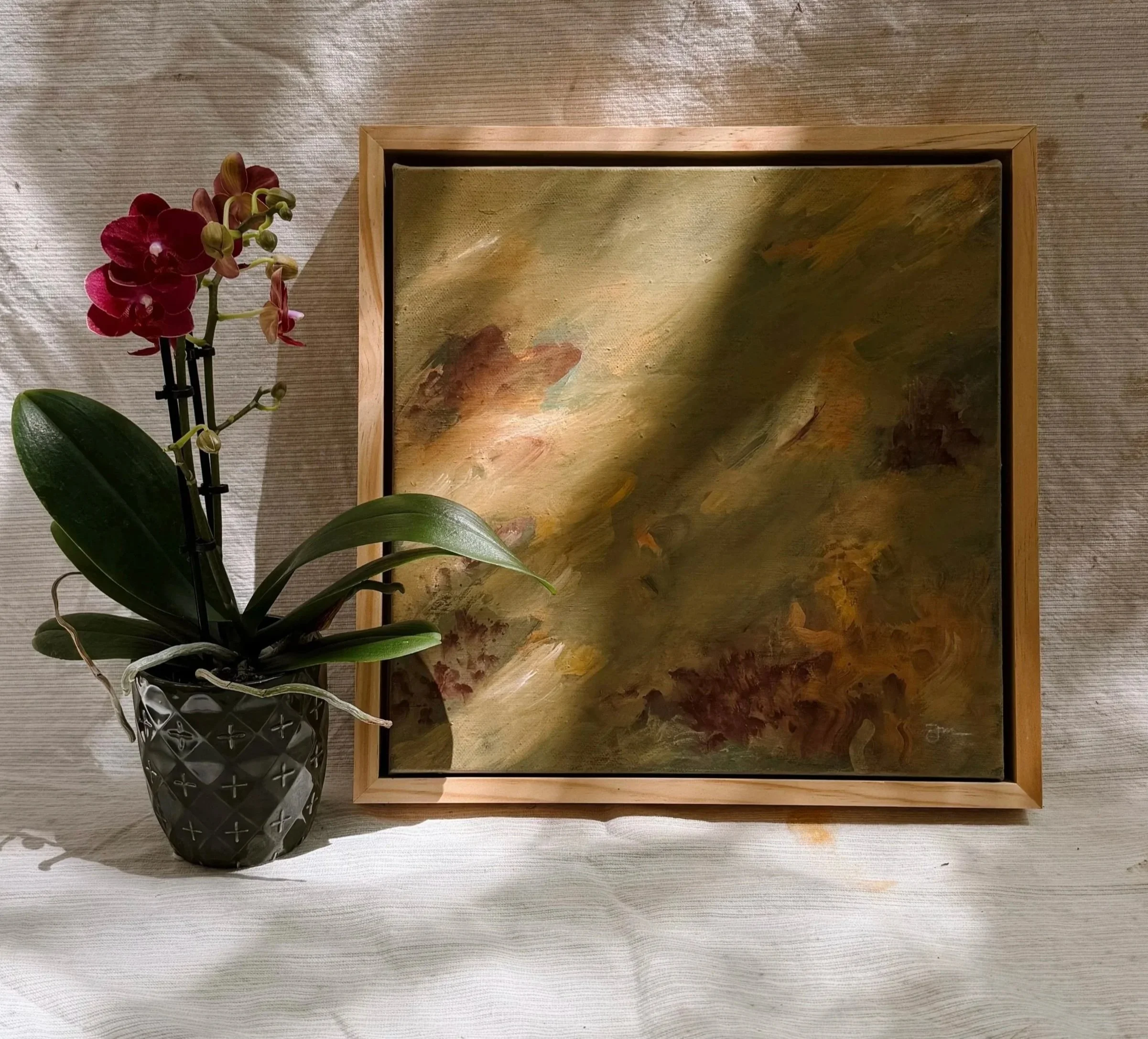 An orchid plant with red flowers and green leaves placed in a decorative dark green pot, next to a framed abstract painting with earthy tones, on a textured canvas surface.