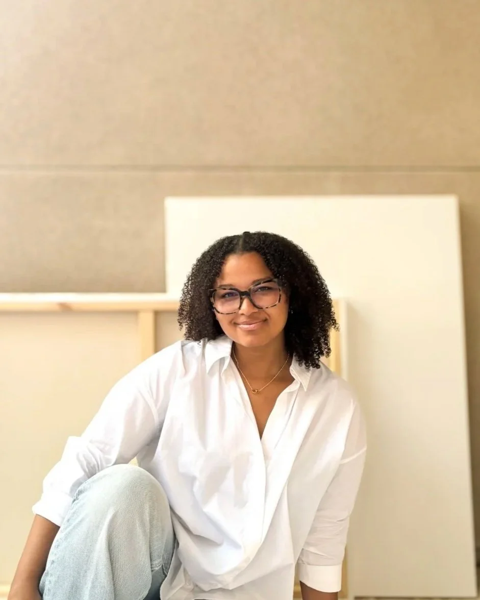 Woman with curly hair and glasses sitting against a neutral background, smiling at the camera.